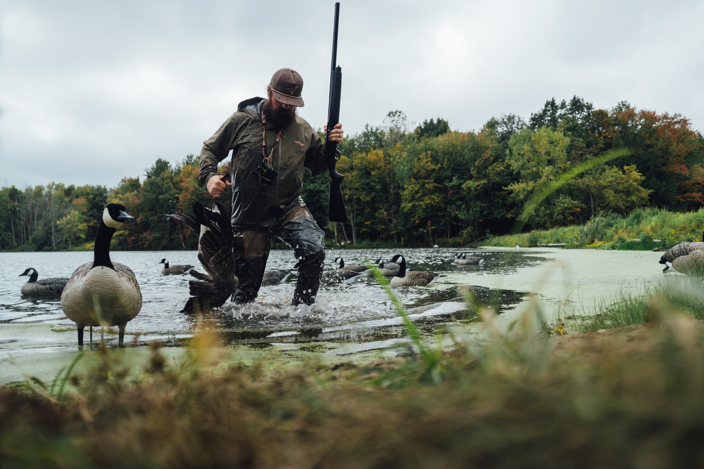Person wearing a hood and backpack stepping out of a boat into shallow water, holding two fishing rods with splashing water around.