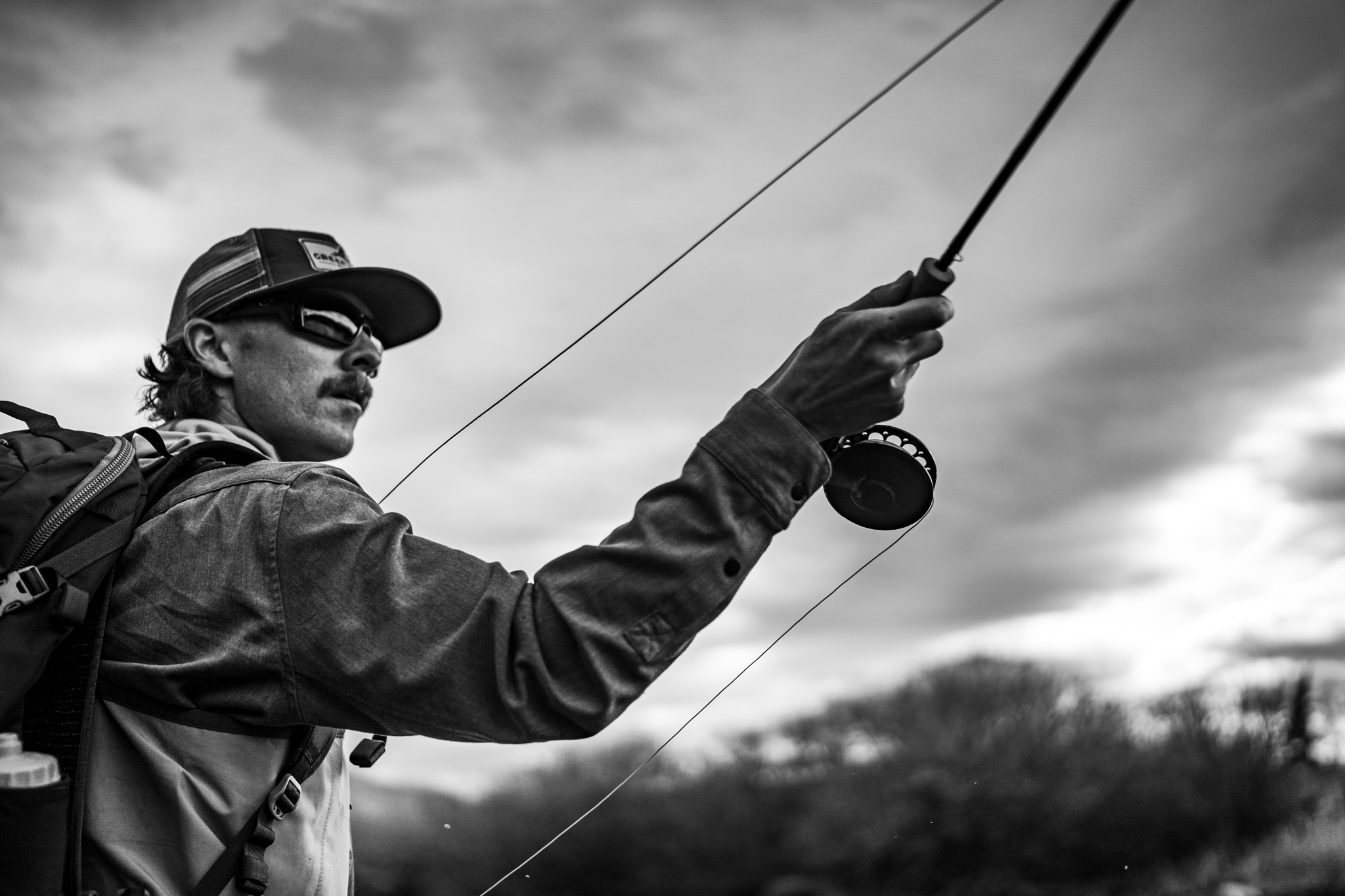 Person wearing a hood and backpack stepping out of a boat into shallow water, holding two fishing rods with splashing water around.