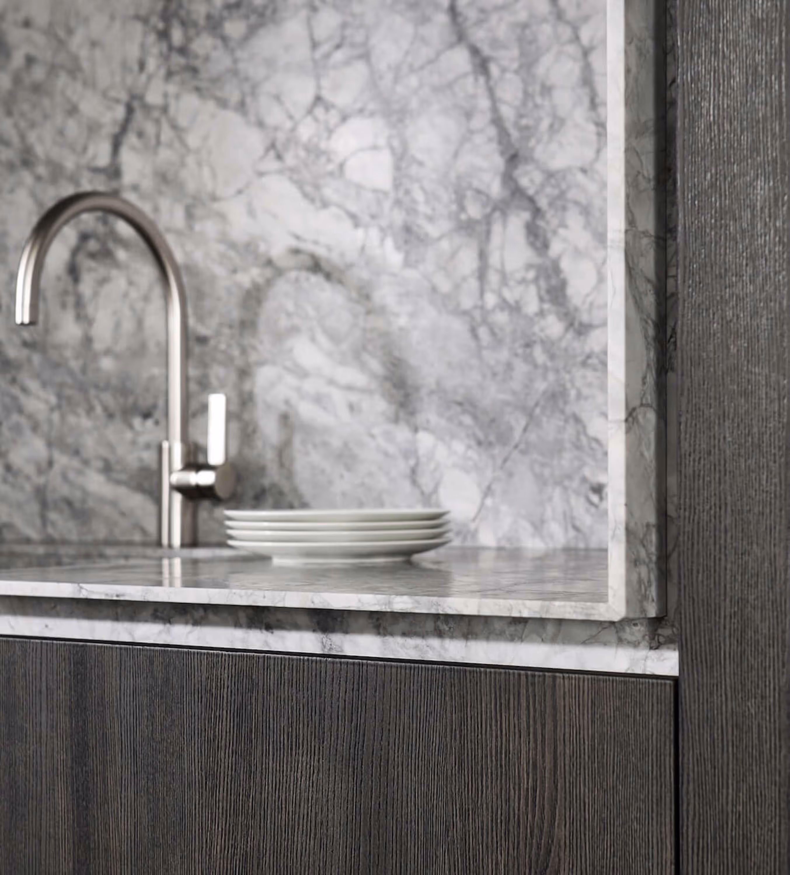 Modern kitchen sink area with a stainless steel faucet, stacked white plates on a gray marble countertop and backsplash, and dark wood cabinetry.