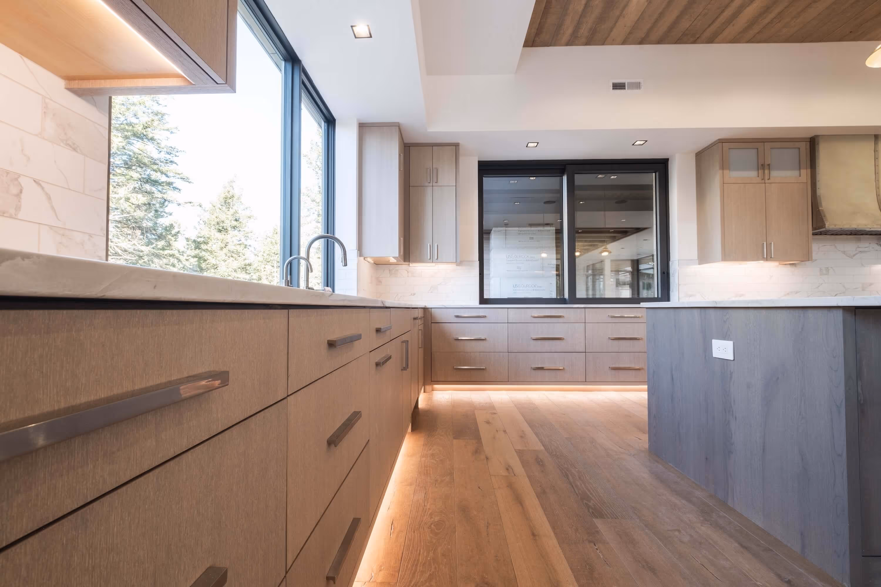 Modern kitchen with wooden cabinets, large windows, marble countertops, and recessed lighting.