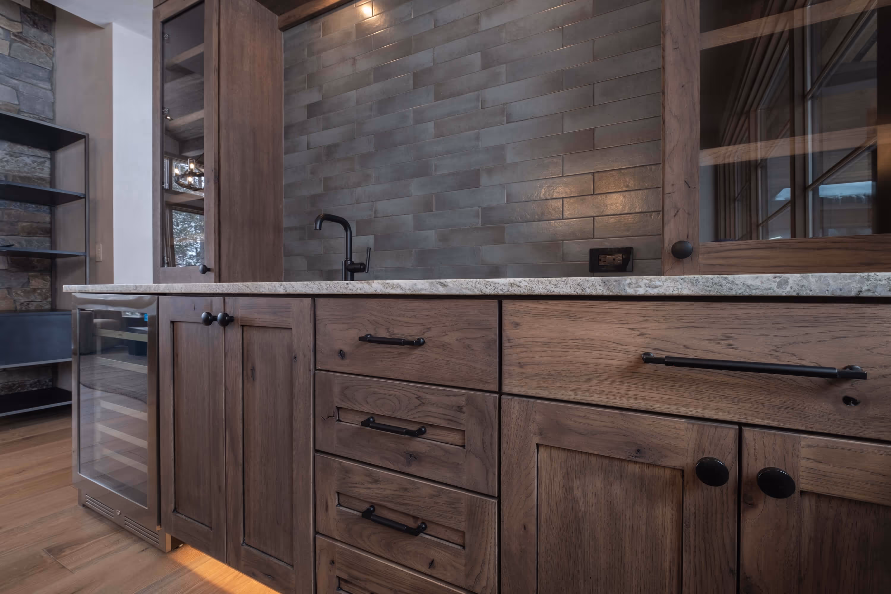 Close-up of wooden kitchen cabinets with black handles, a marble countertop, a black faucet, and a tiled gray backsplash.