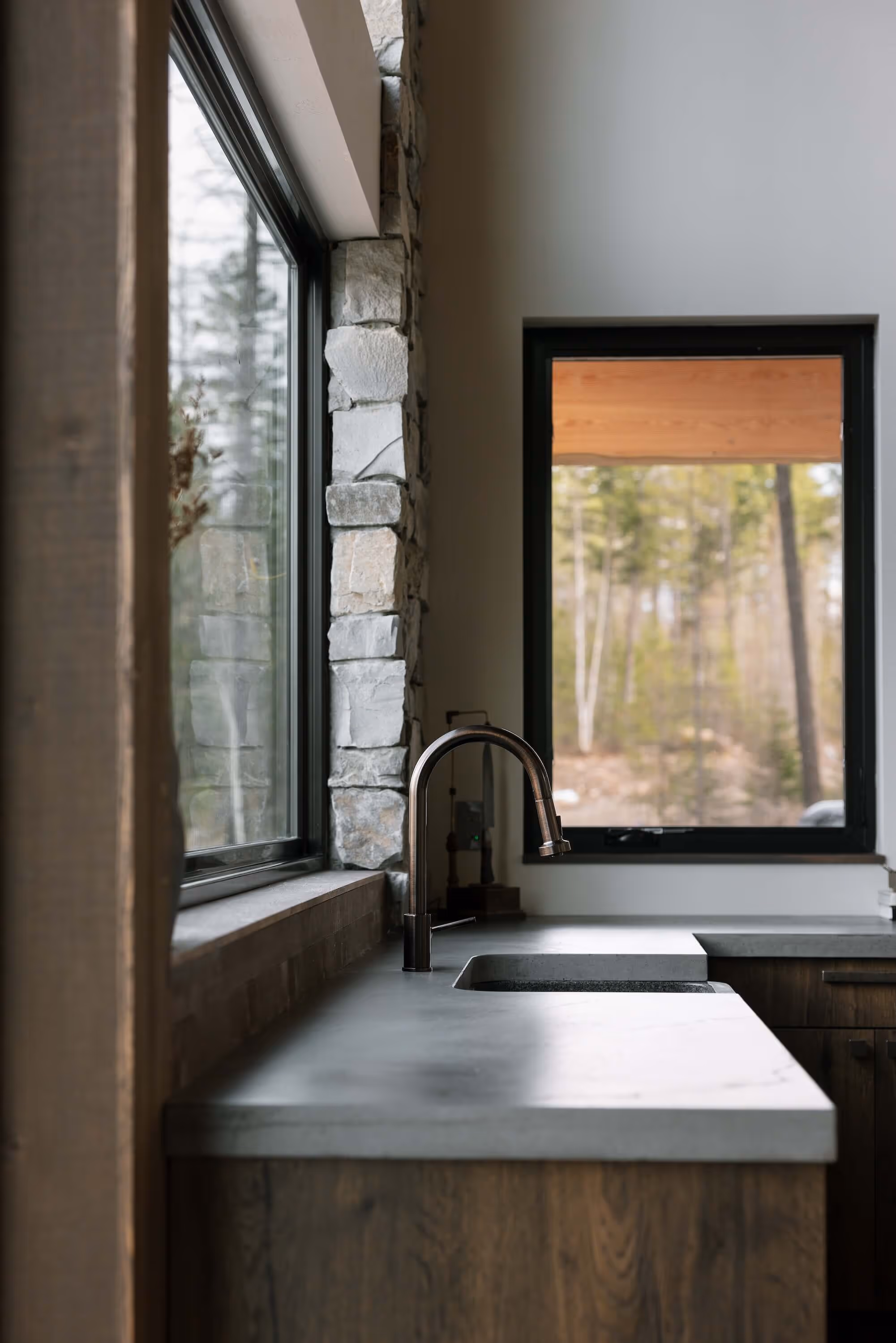 Modern kitchen sink with a curved faucet set in a concrete countertop beside large windows showing an outdoor wooded scene.