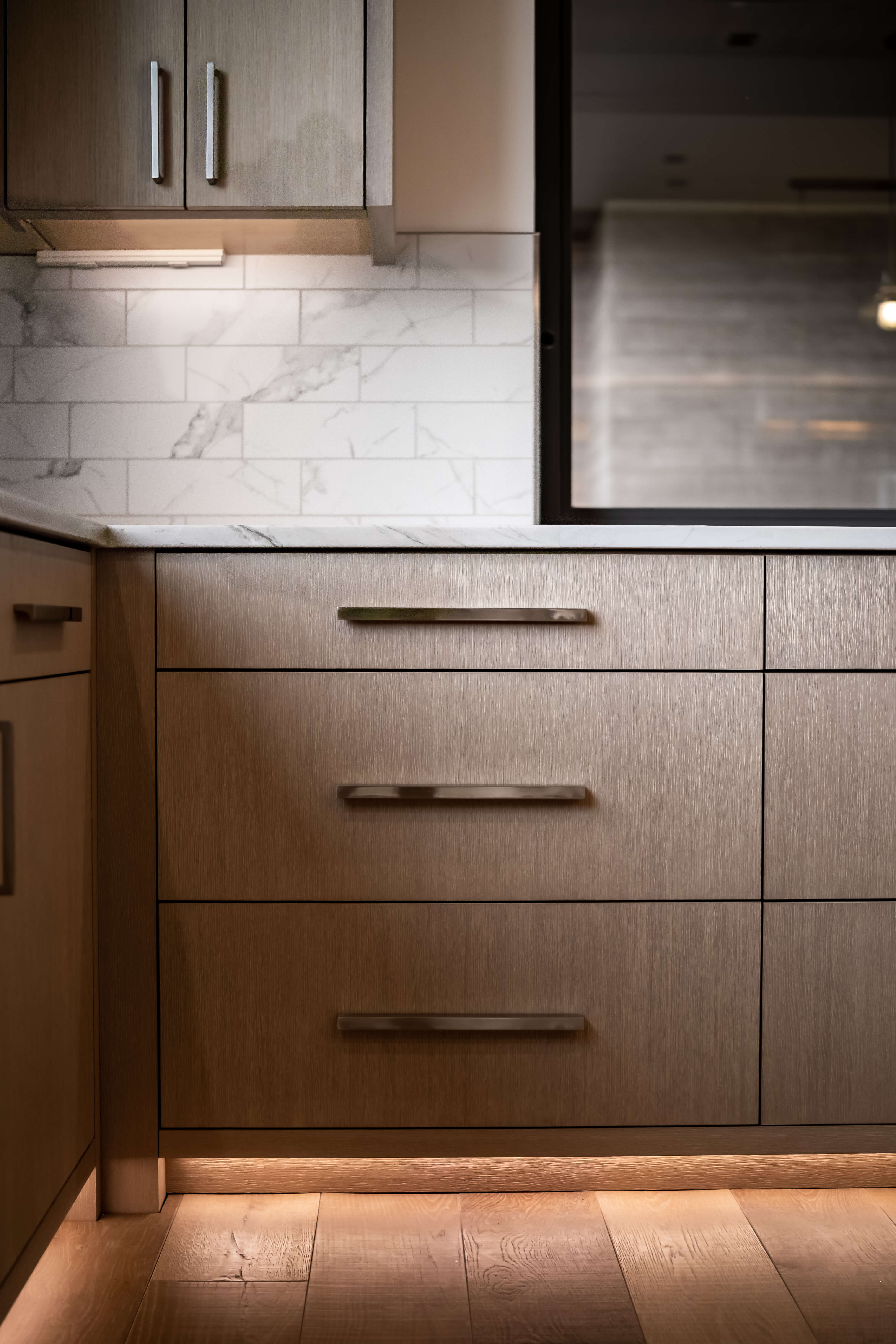 Modern kitchen with blue cabinetry, wooden upper cabinets with glass doors, a gray marble island, stainless steel stove, and large windows overlooking trees.