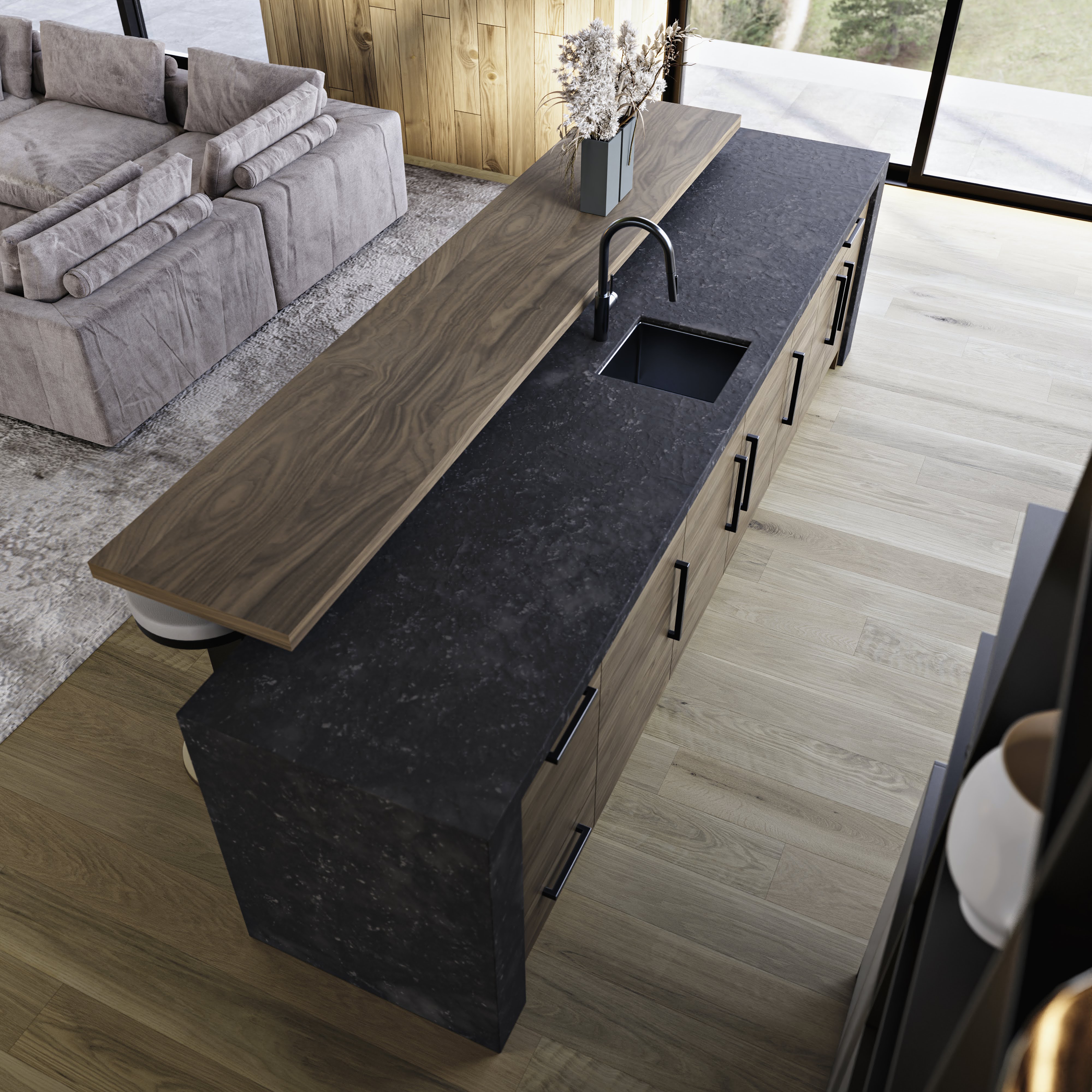 Modern kitchen island with black stone countertop, built-in sink, wooden bar counter, and light wood flooring next to gray sectional sofa.