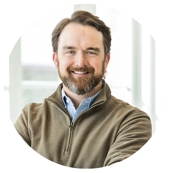 A beared man smiling wearing a brown quaterzip pullover over a light blue collared shirt