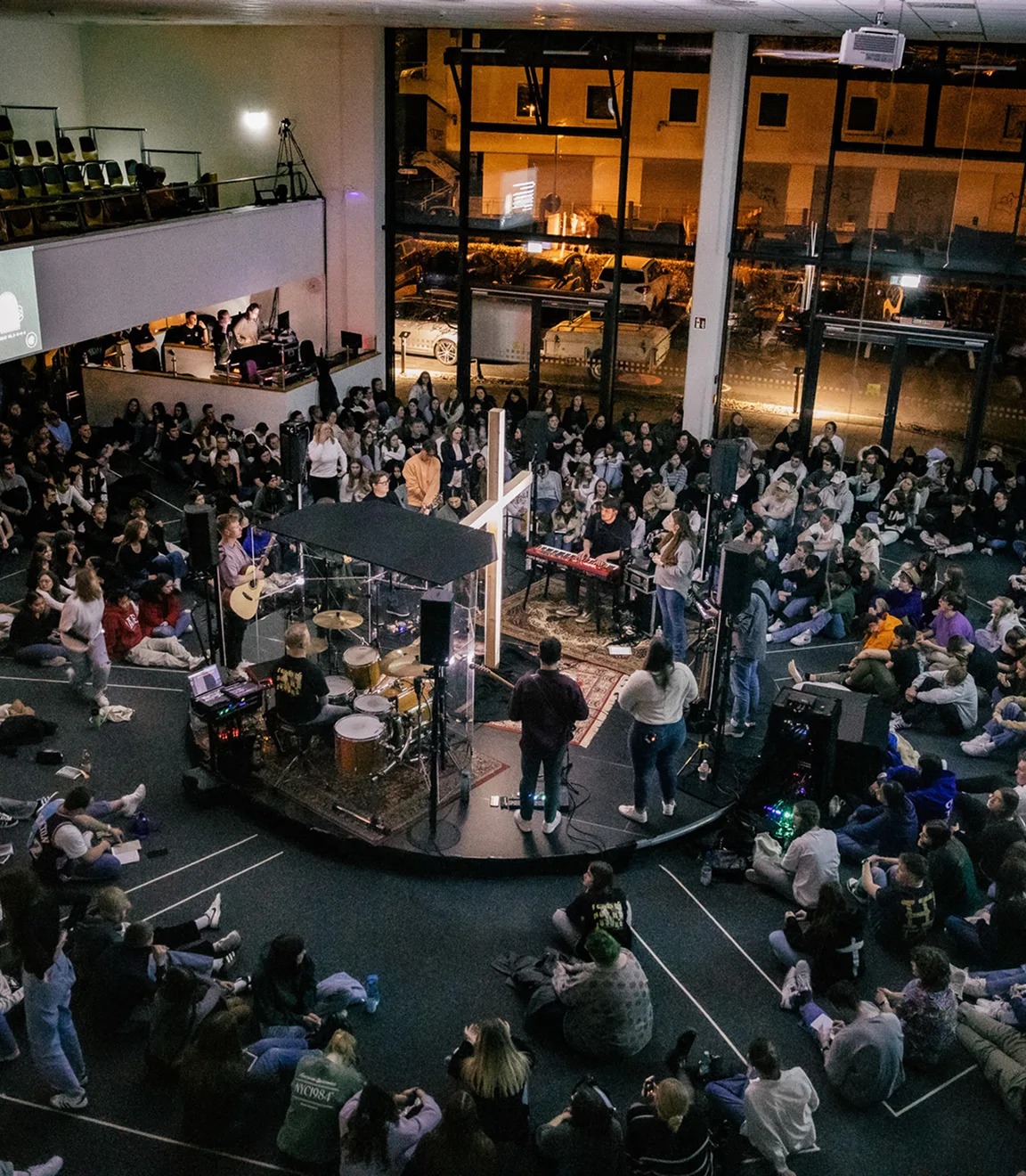 Large group of people seated on the floor around a circular stage with musicians playing instruments and a large illuminated cross in the center.