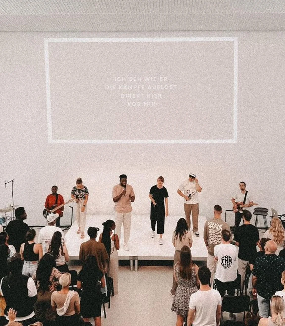 A diverse group of people standing and facing a stage where a band and singers perform in a minimalist white room.