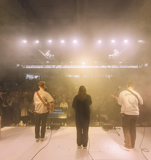 View from stage showing three musicians performing before a large, dimly lit audience under bright stage lights.