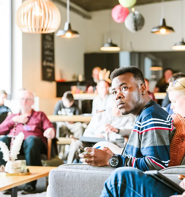 Man in a striped sweater attentively listening in a casual group meeting with people seated around tables in a bright room.