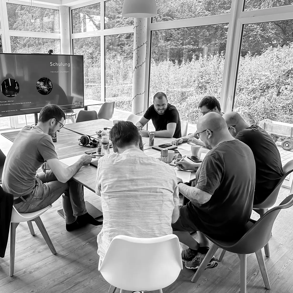 Six people sitting around a table in a bright room with large windows, engaged in discussion and writing, with a screen displaying a presentation titled 'Schulung'.