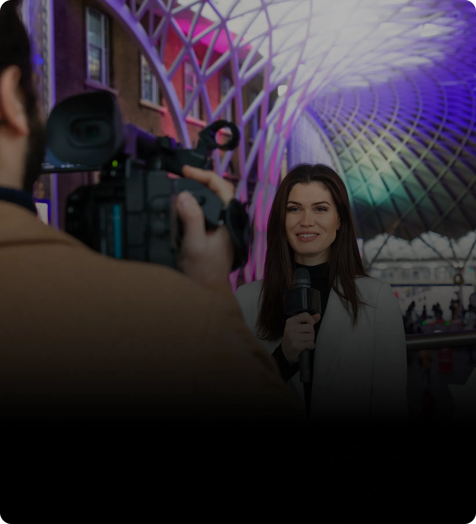 Female reporter holding a microphone and smiling at the camera with a colorful, arched glass ceiling in the background.