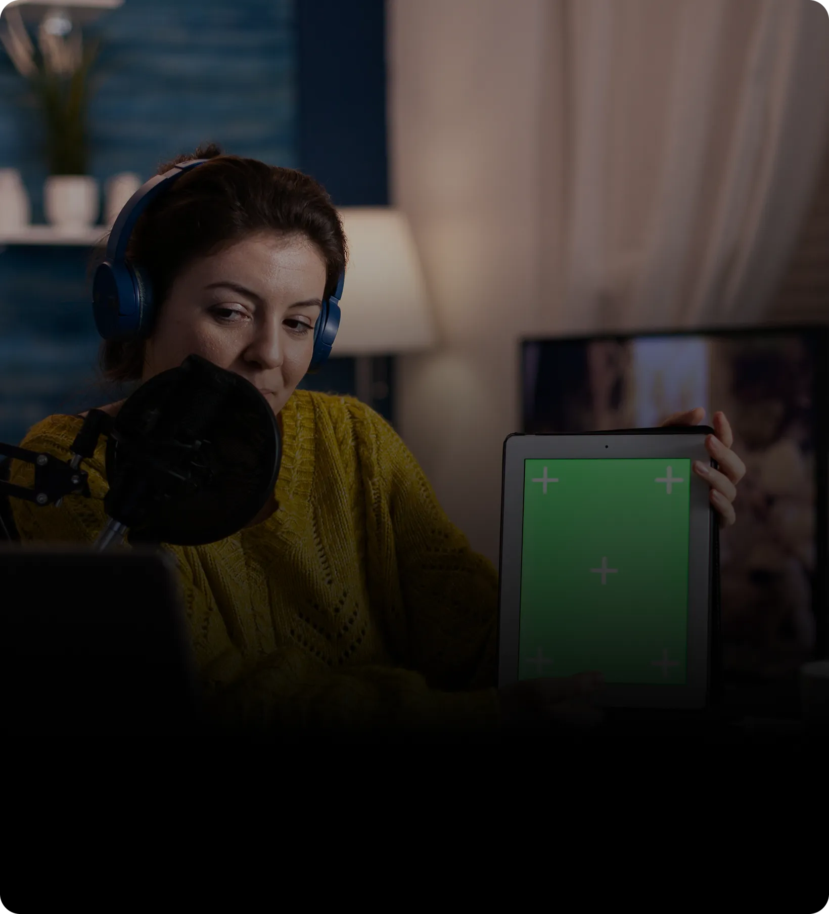 Woman in blue headphones speaking into a microphone while holding a tablet with a green screen in a cozy room.