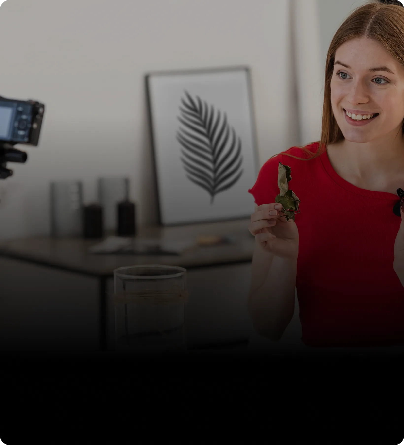 Smiling woman in red shirt holding a dried leaf, sitting in front of a camera indoors.