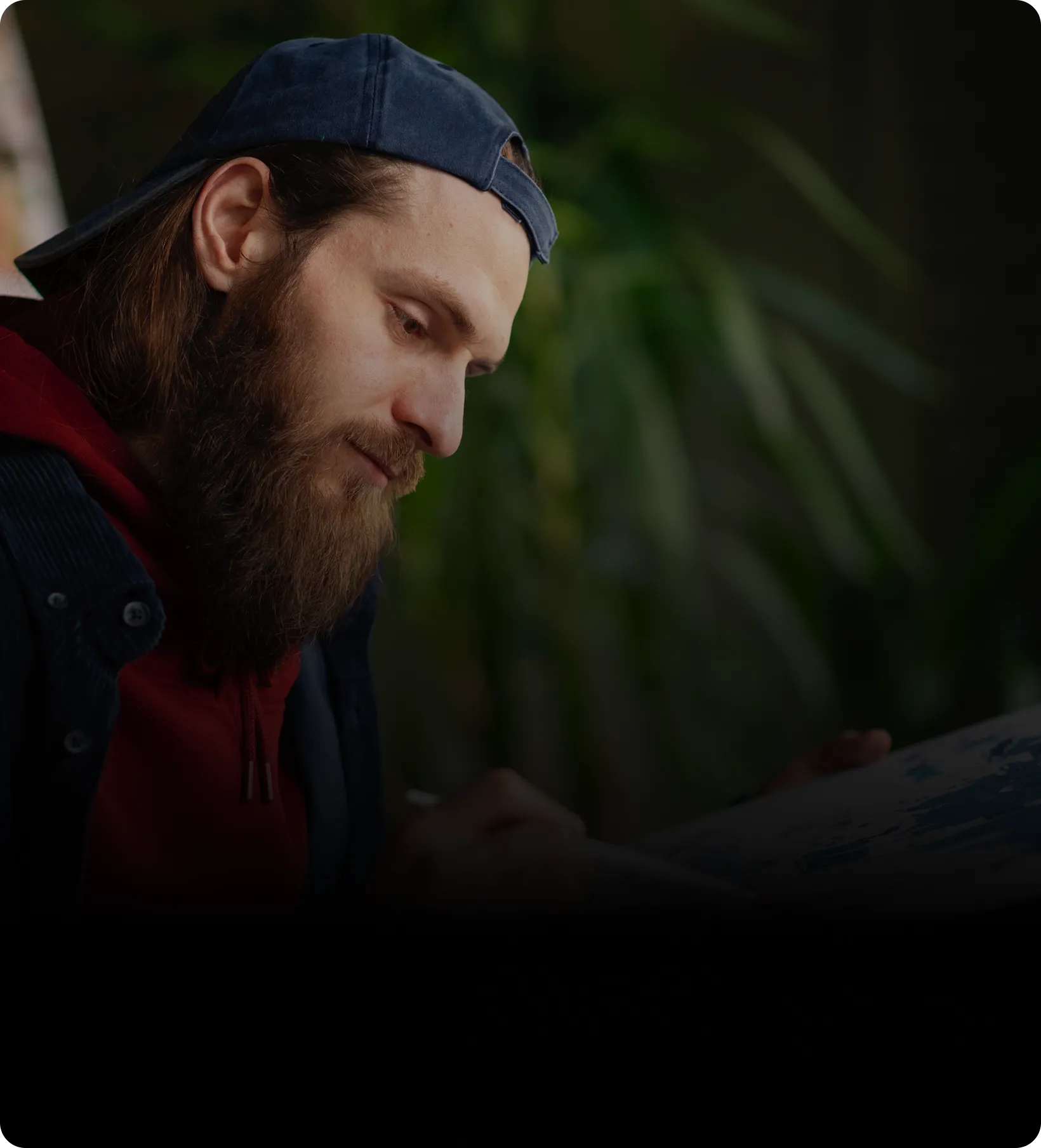 Bearded man wearing a backwards cap and red hoodie focused on painting or drawing.