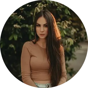 Portrait of a woman with long dark hair, wearing a brown top, standing outdoors with greenery in the background.