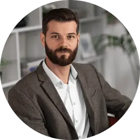 Portrait of a man with a beard wearing a gray blazer and white shirt sitting indoors.