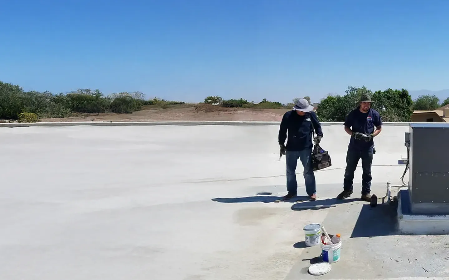 Workers apply a waterproof coating on a rooftop under clear blue skies.