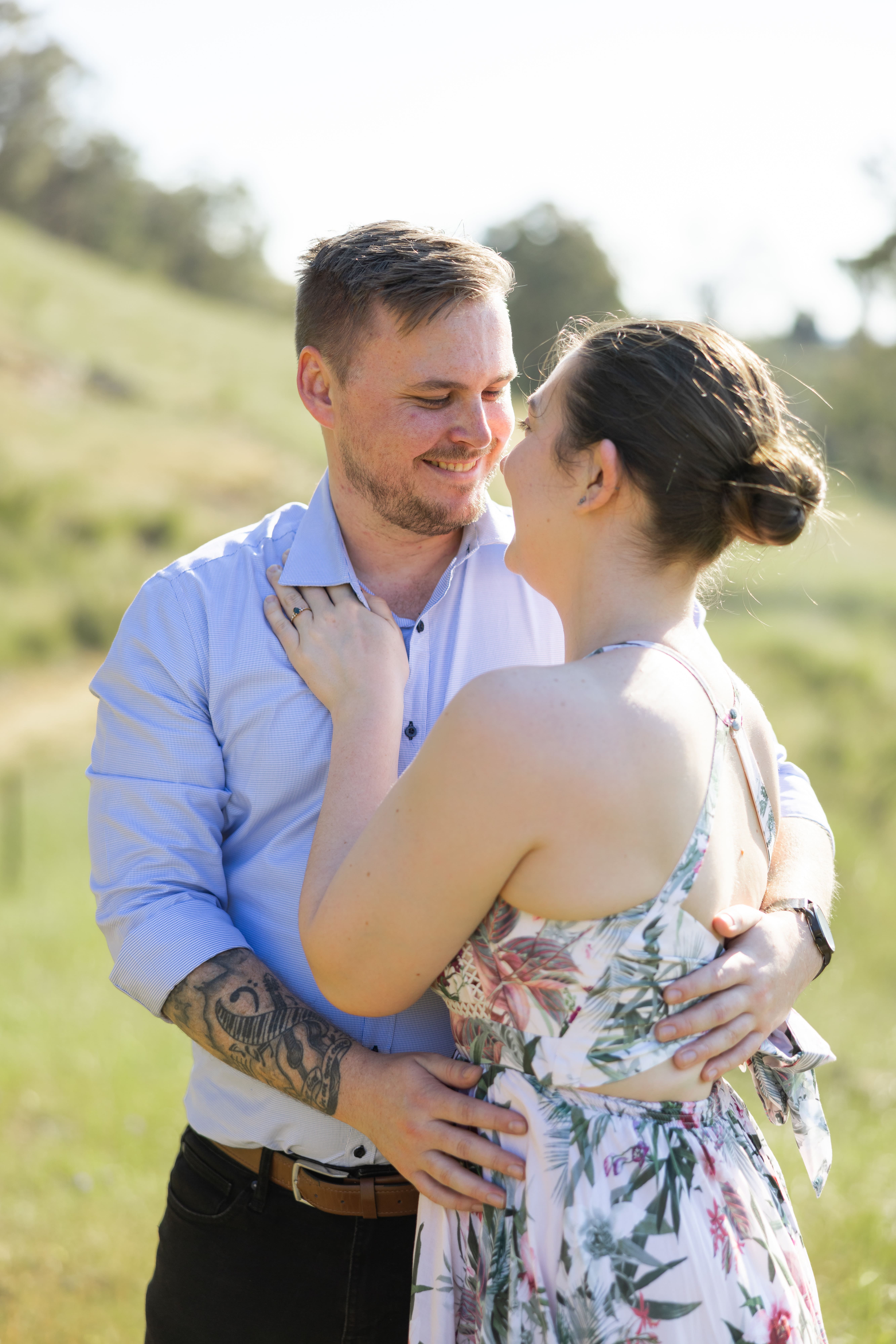 Smiling couple outdoors sitting on grass with greenery and hills in the background, the woman wearing a floral dress and the man in a light blue shirt.