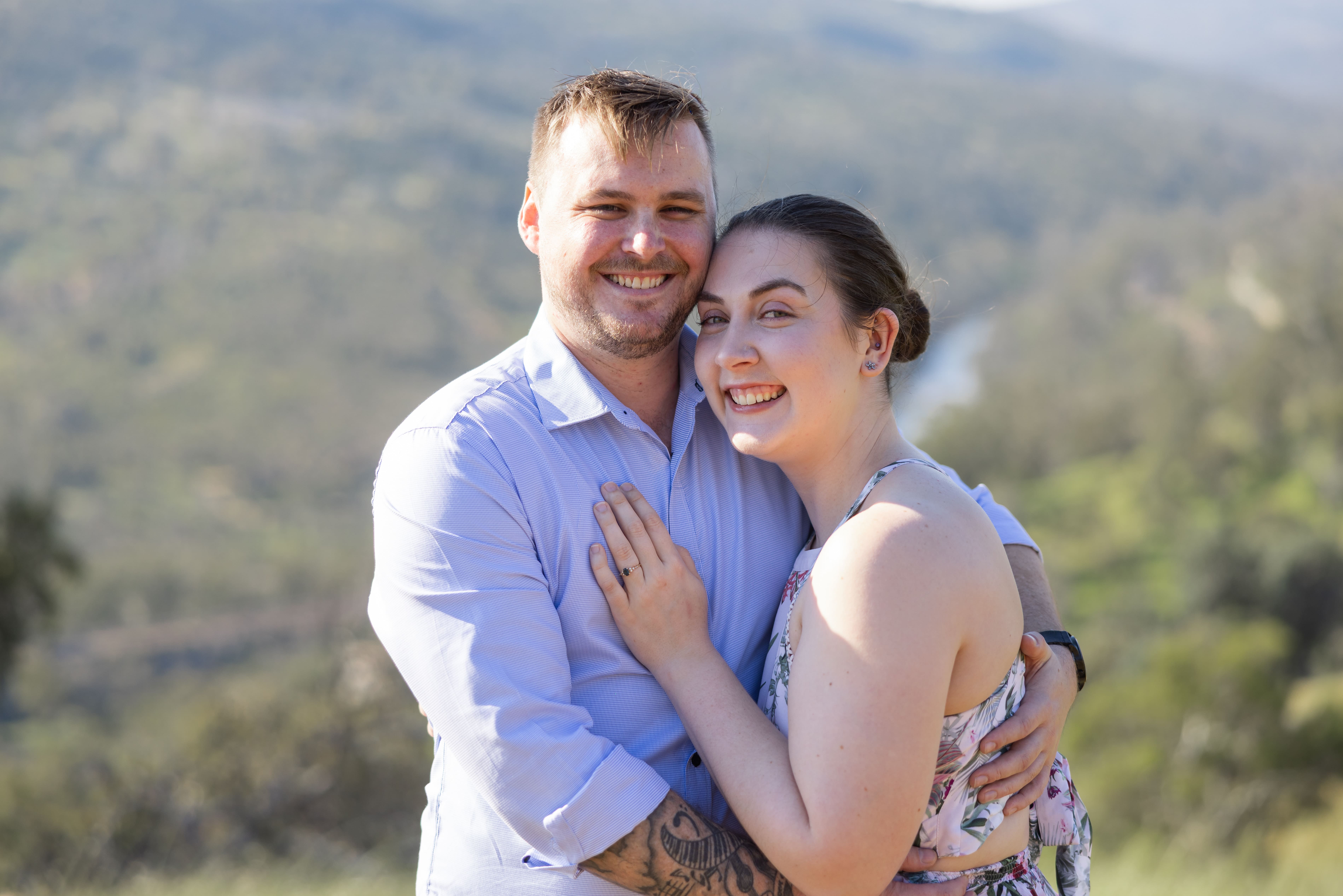Smiling couple embracing outdoors with scenic green hills and river in background.