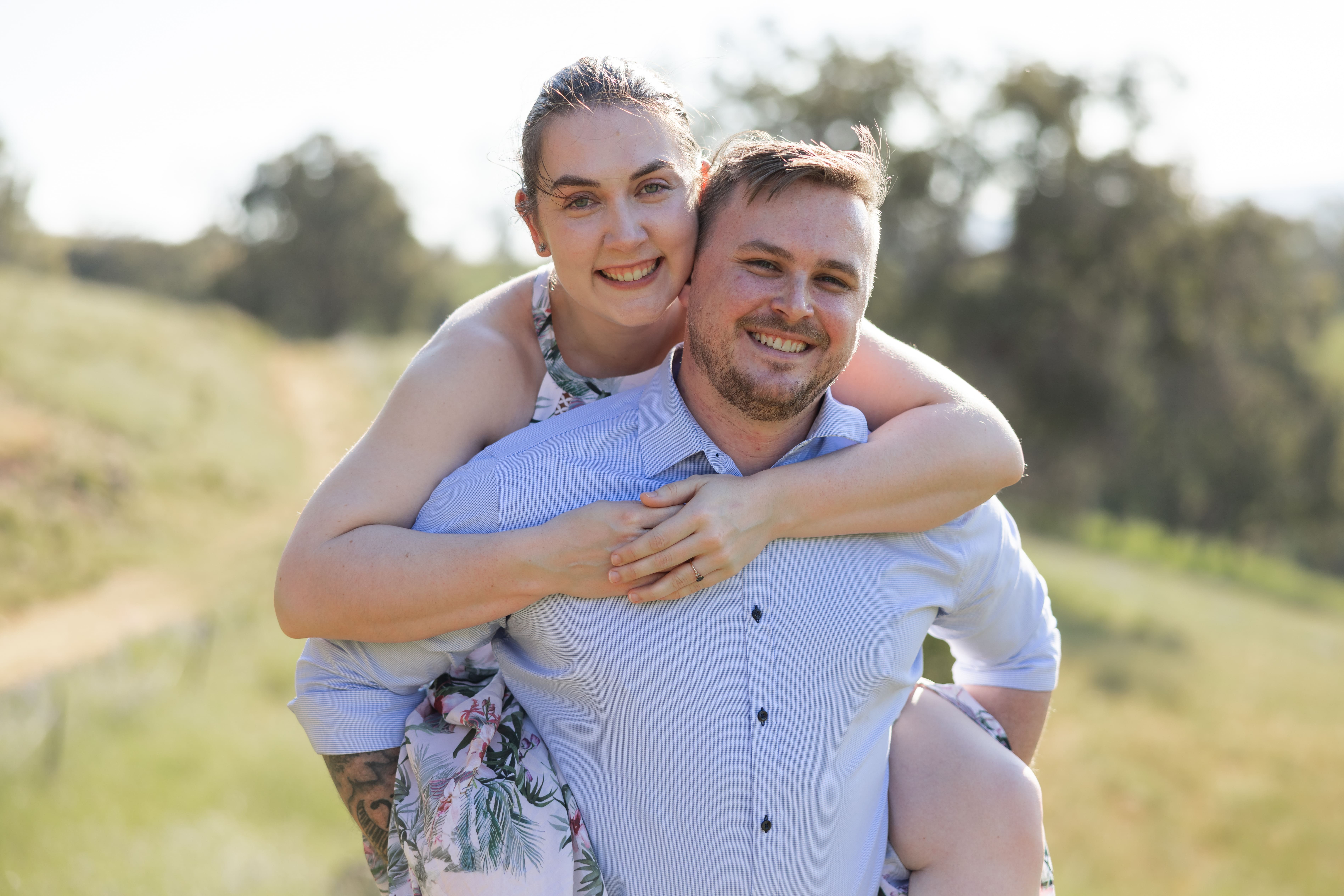 Smiling couple outdoors sitting on grass with greenery and hills in the background, the woman wearing a floral dress and the man in a light blue shirt.