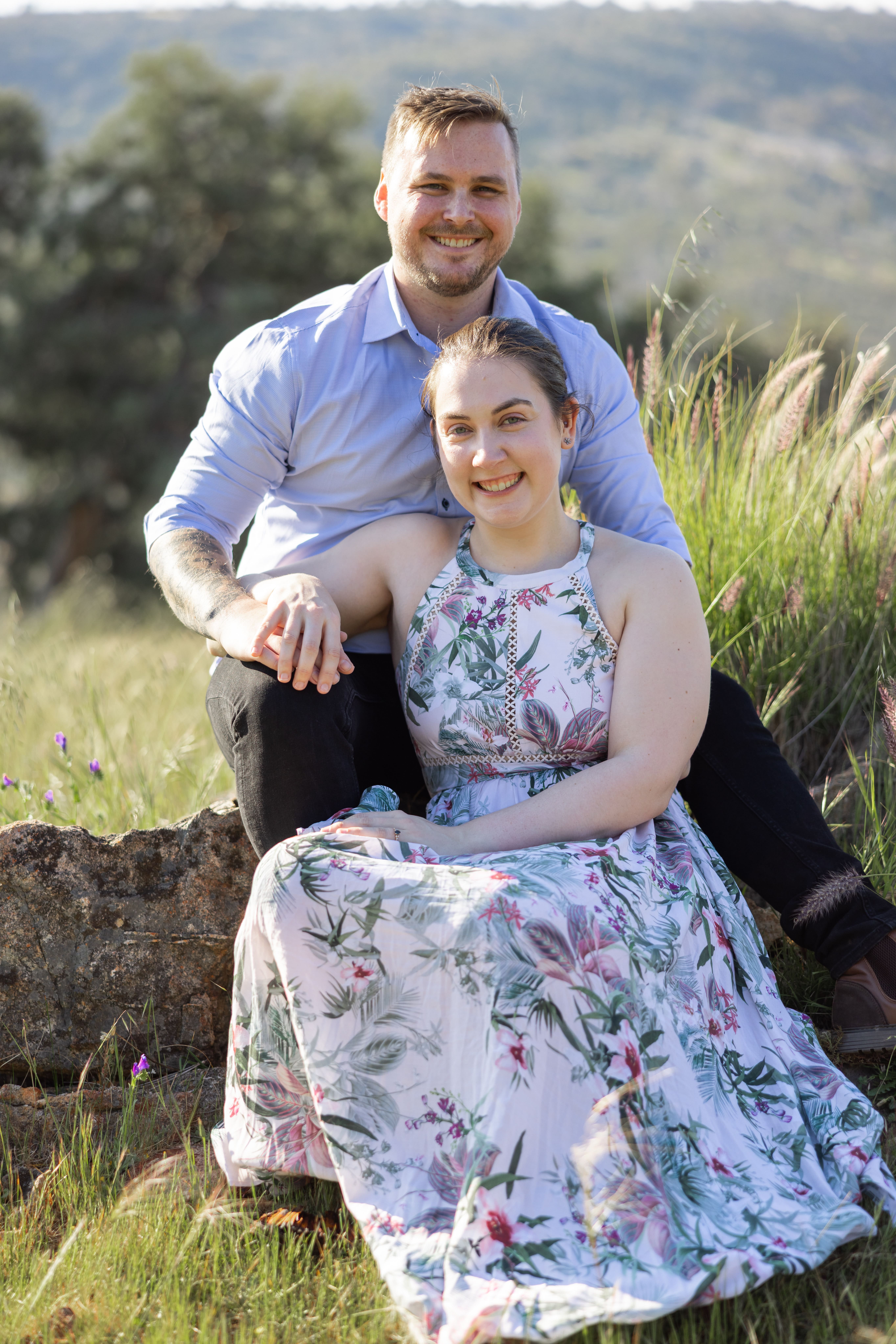 Smiling couple outdoors sitting on grass with greenery and hills in the background, the woman wearing a floral dress and the man in a light blue shirt.