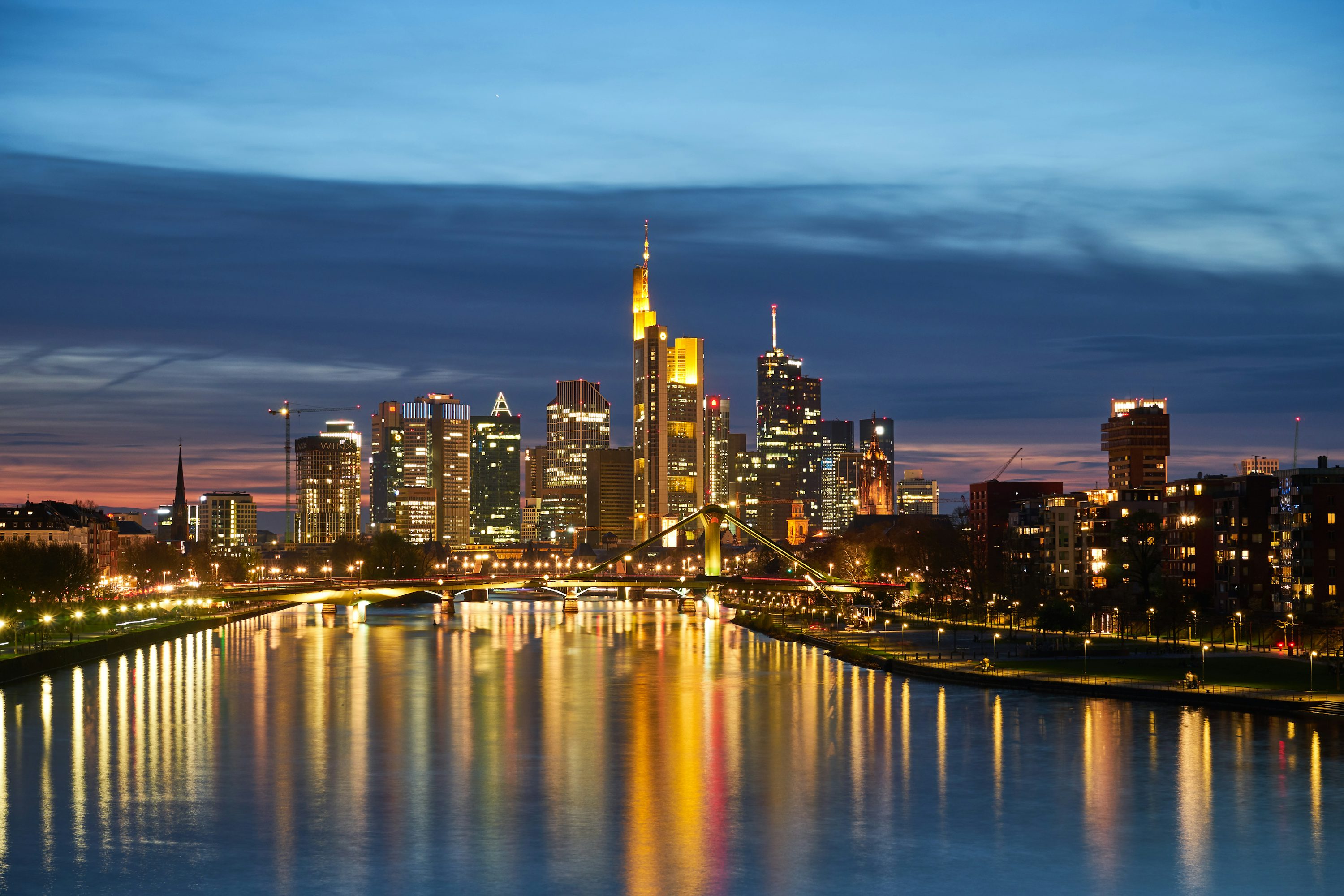 Frankfurt city skyline at dusk with illuminated skyscrapers reflecting on the river and a bridge in the foreground.