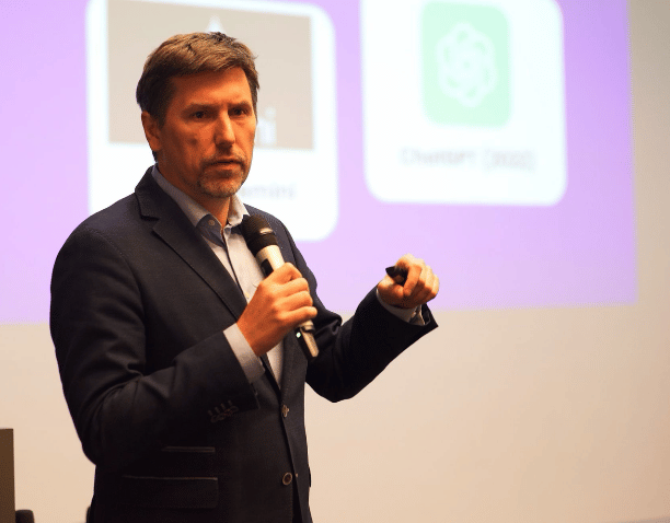 dr Paweł Kowalski w marynarce trzyma mikrofon i prowadzi prezentację z widocznym ekranem w tle.