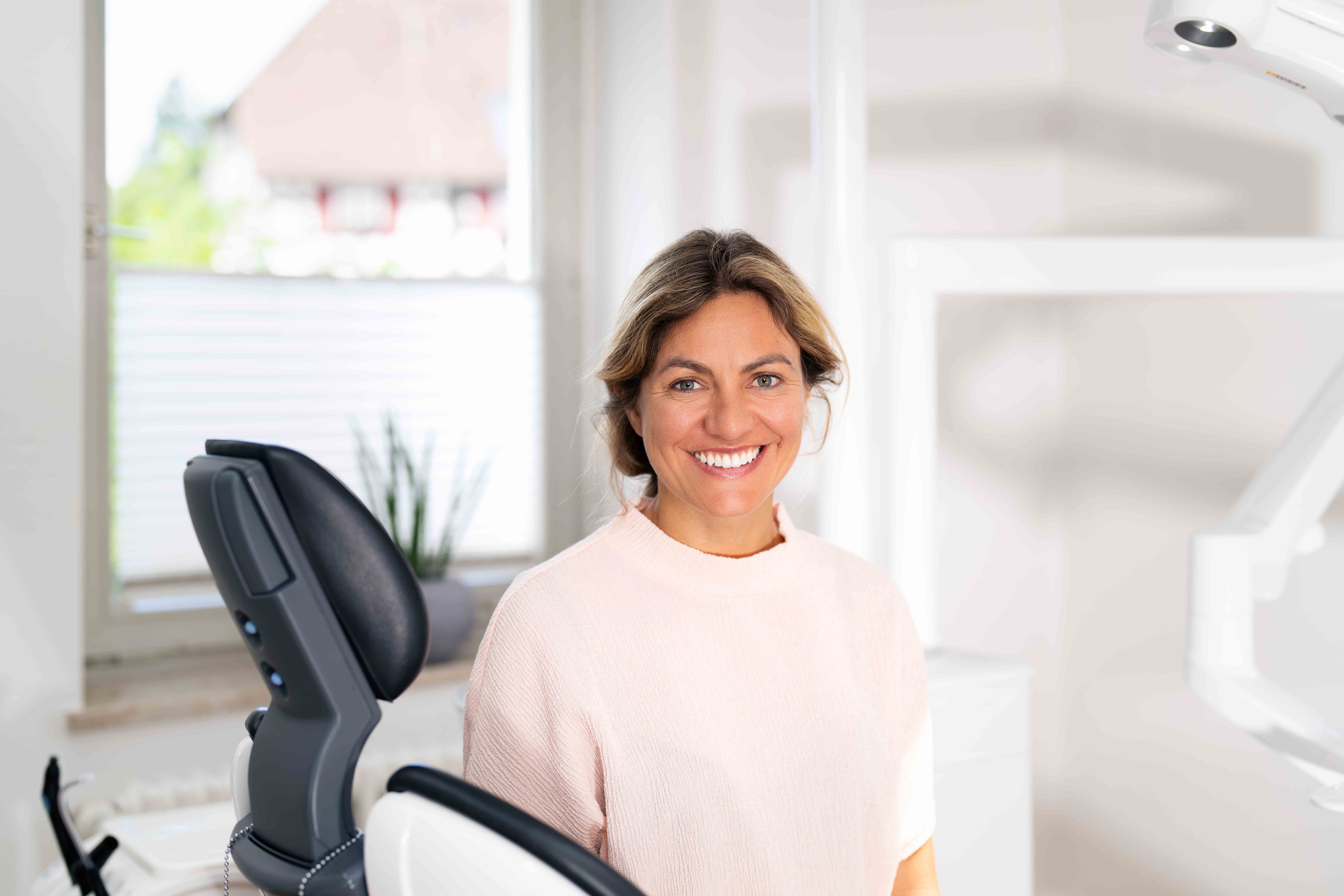 Smiling woman in light pink sweater sitting in a modern dental office next to a dental chair.