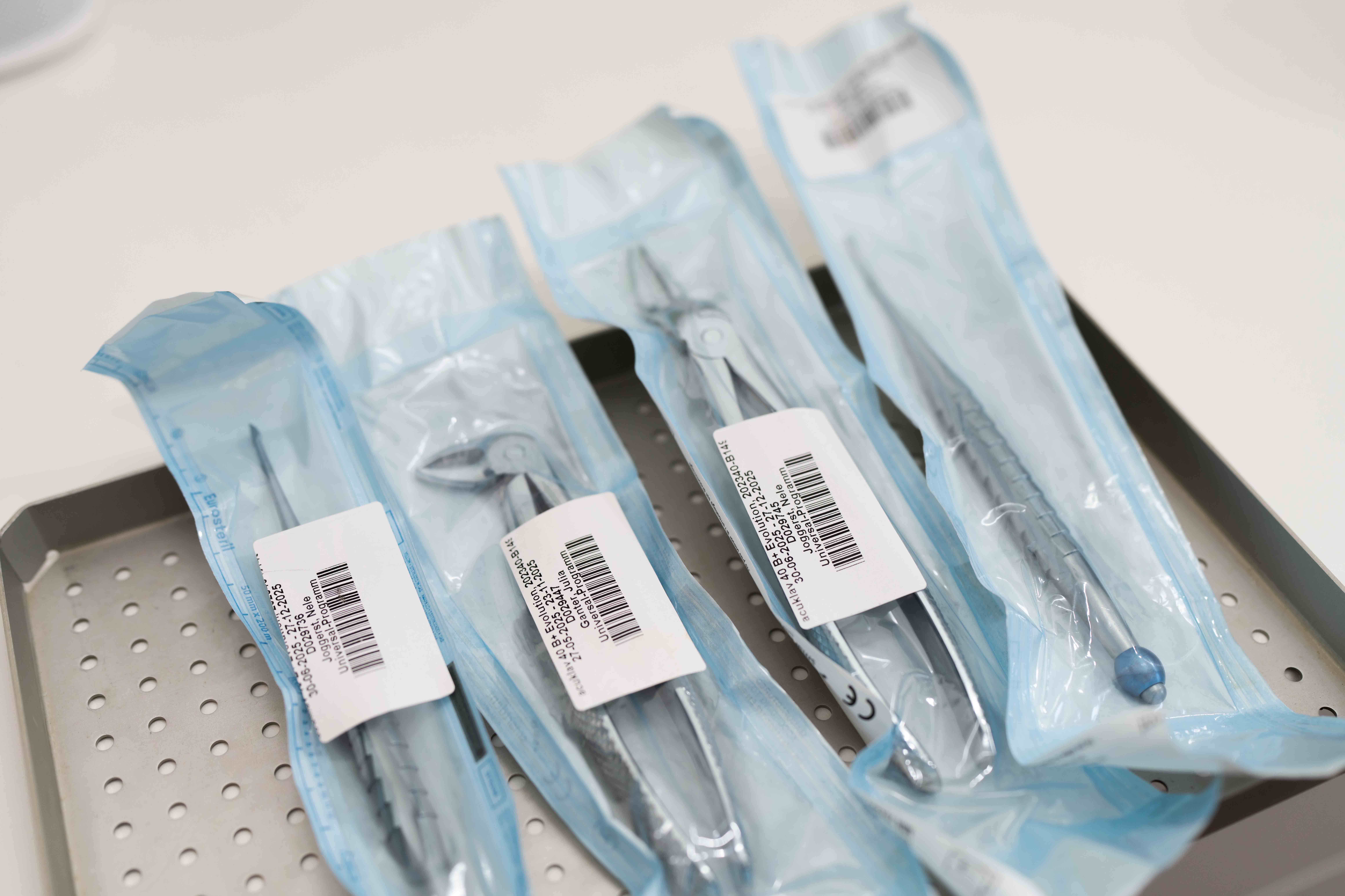 Four sterilized dental or surgical instruments in sealed blue packaging on a perforated metal tray.