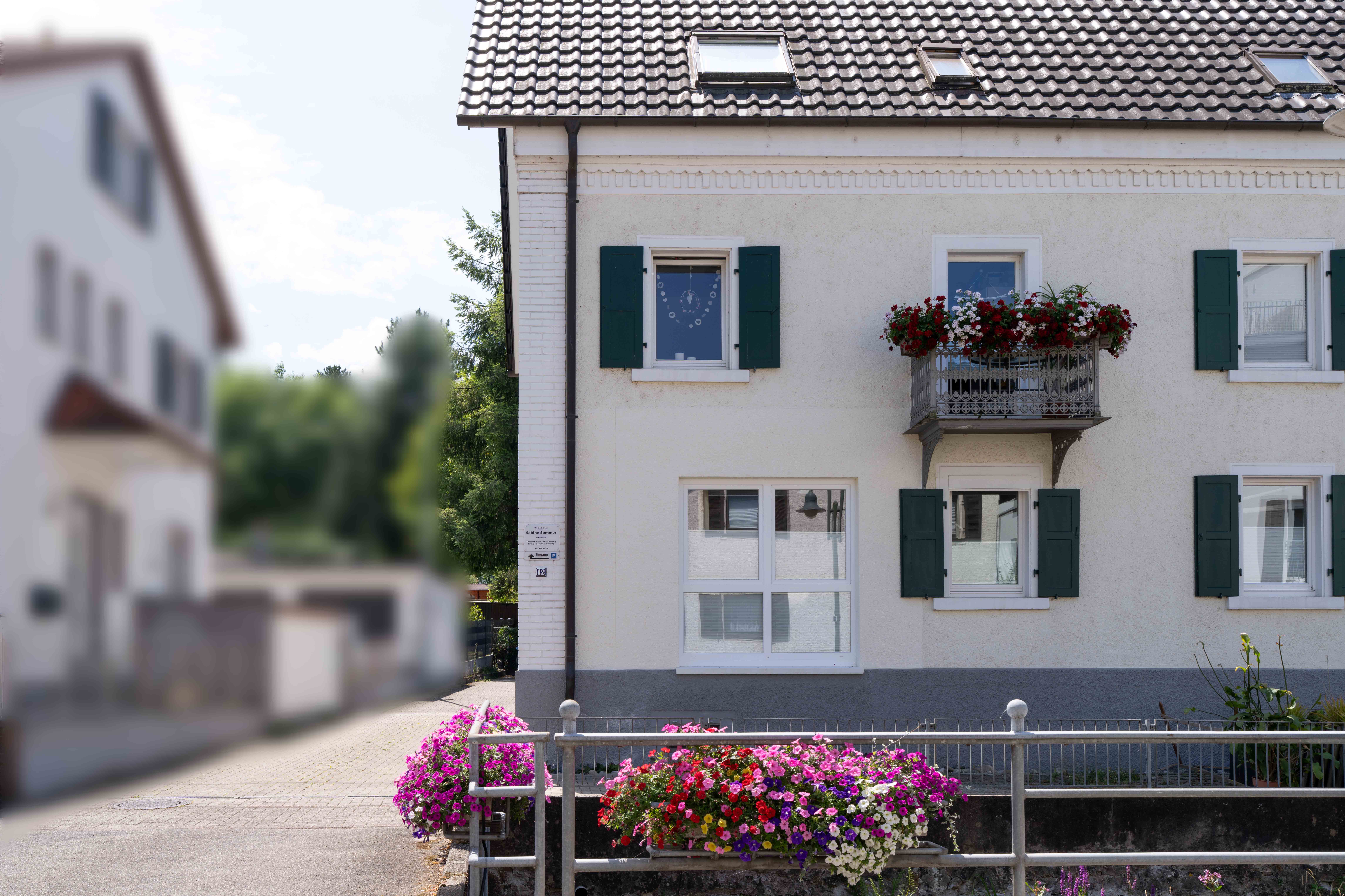 White house facade with green shutters, a balcony with red and white flowers, and a flower bed with colorful blooms in front.