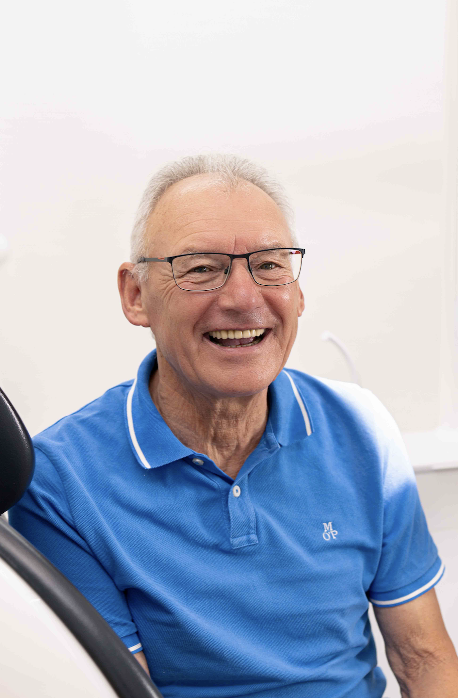 Smiling elderly man wearing glasses and a blue polo shirt sitting in a dental chair.