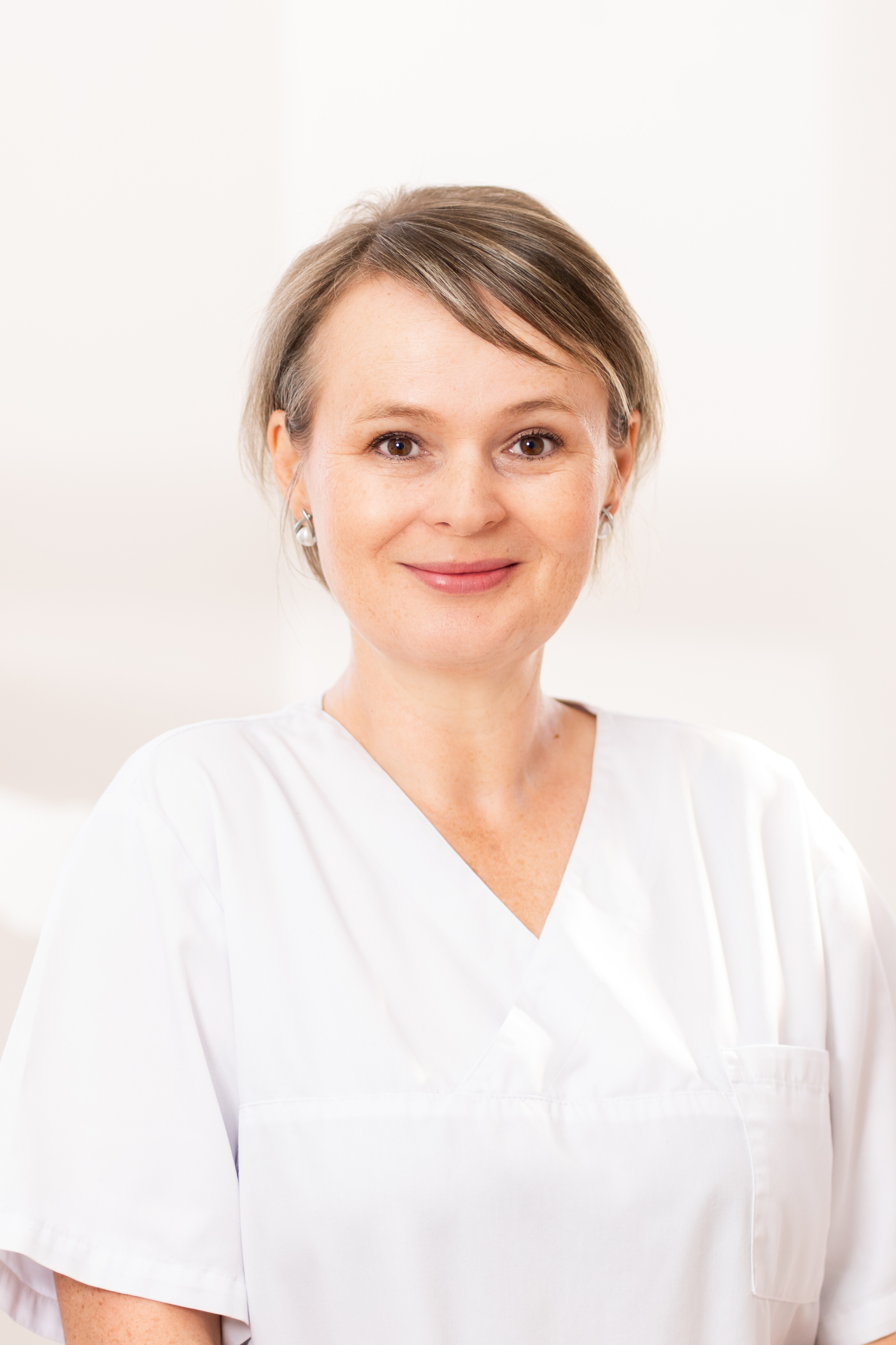 Smiling woman with short hair wearing white medical scrubs in a bright room.