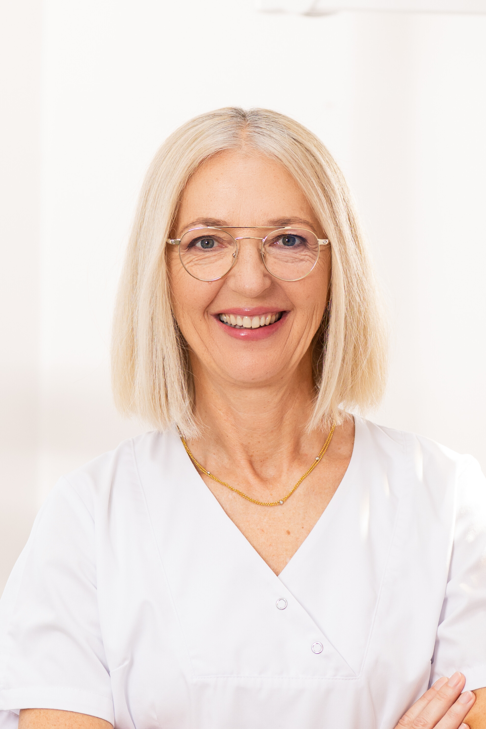 Smiling middle-aged woman with blonde hair and glasses wearing a white medical uniform and a gold necklace.