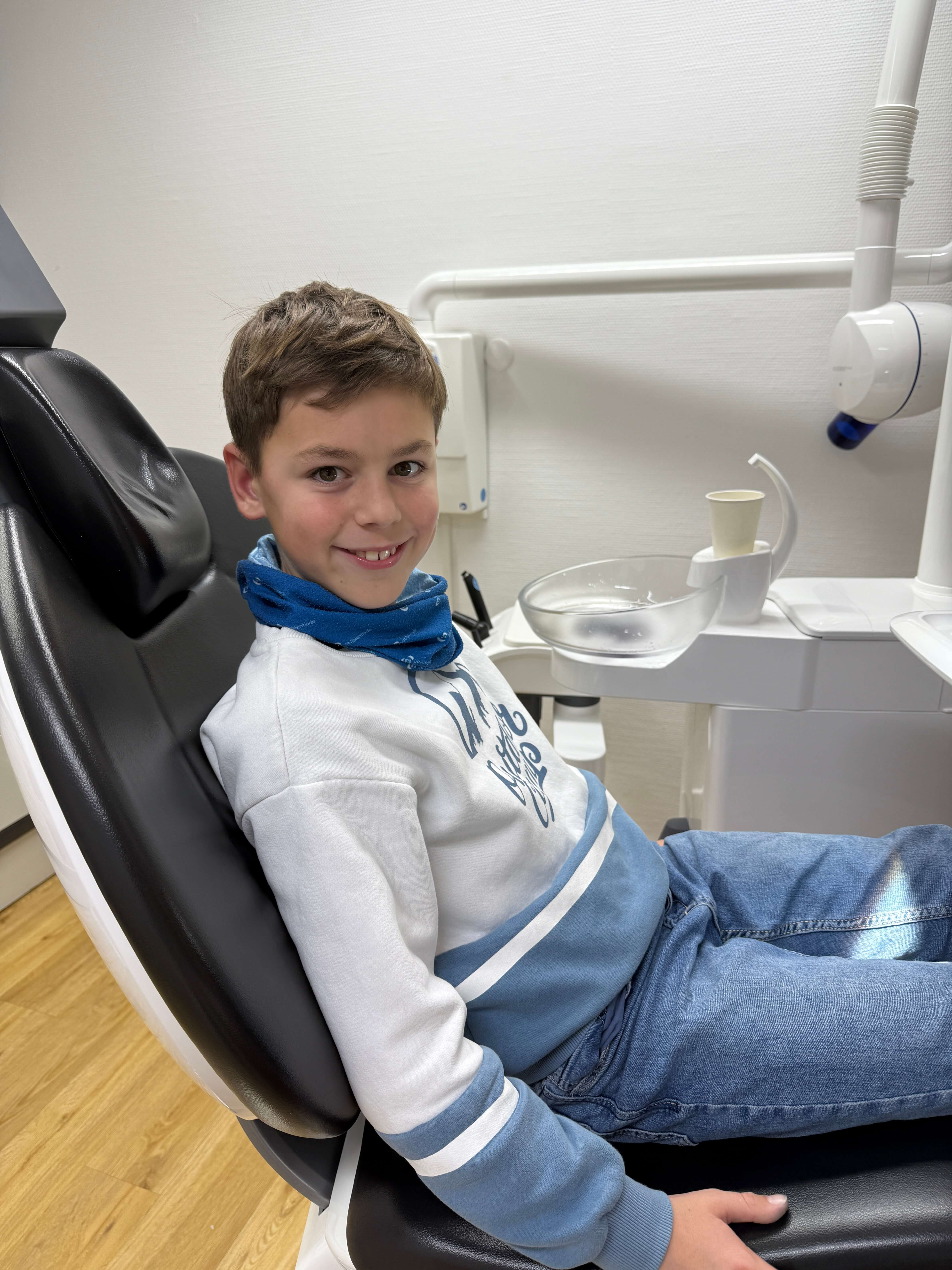 Smiling boy sitting in a dental chair wearing a white and blue sweatshirt and blue jeans.