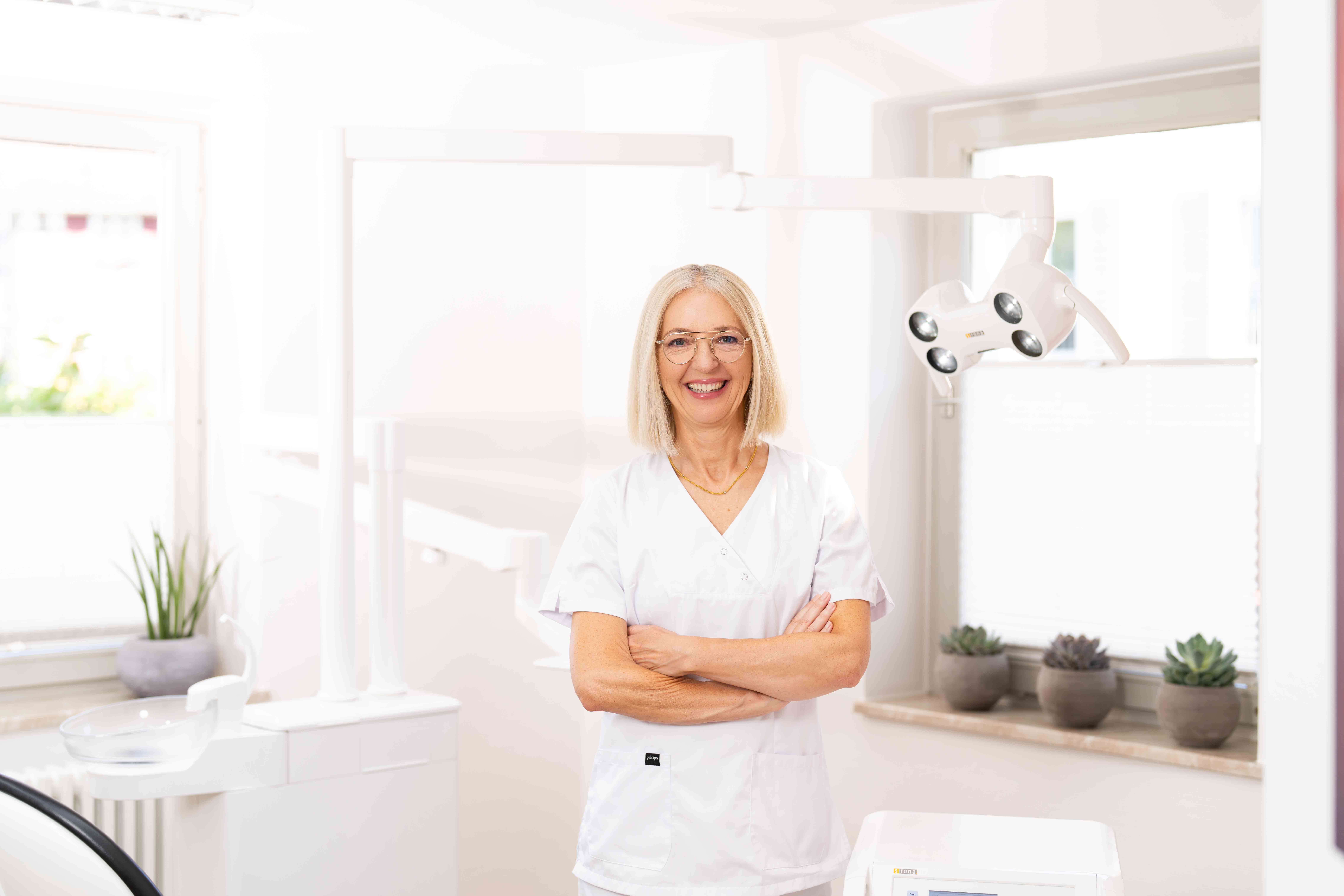 Smiling female dentist in white coat standing in a bright dental clinic with dental equipment and potted plants in the background.
