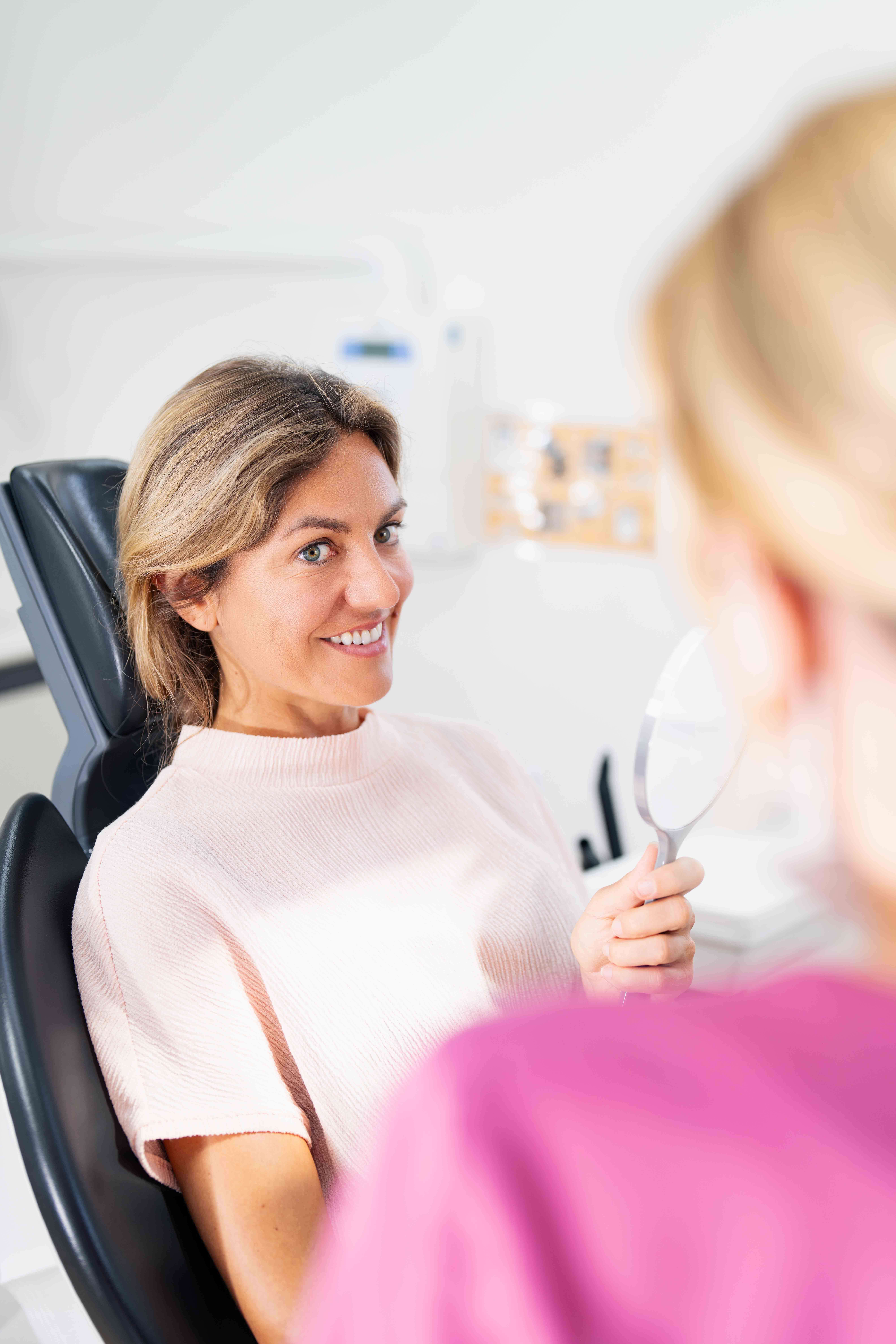 Woman in a dental chair smiling and looking at herself in a handheld mirror held by a person in a pink top.