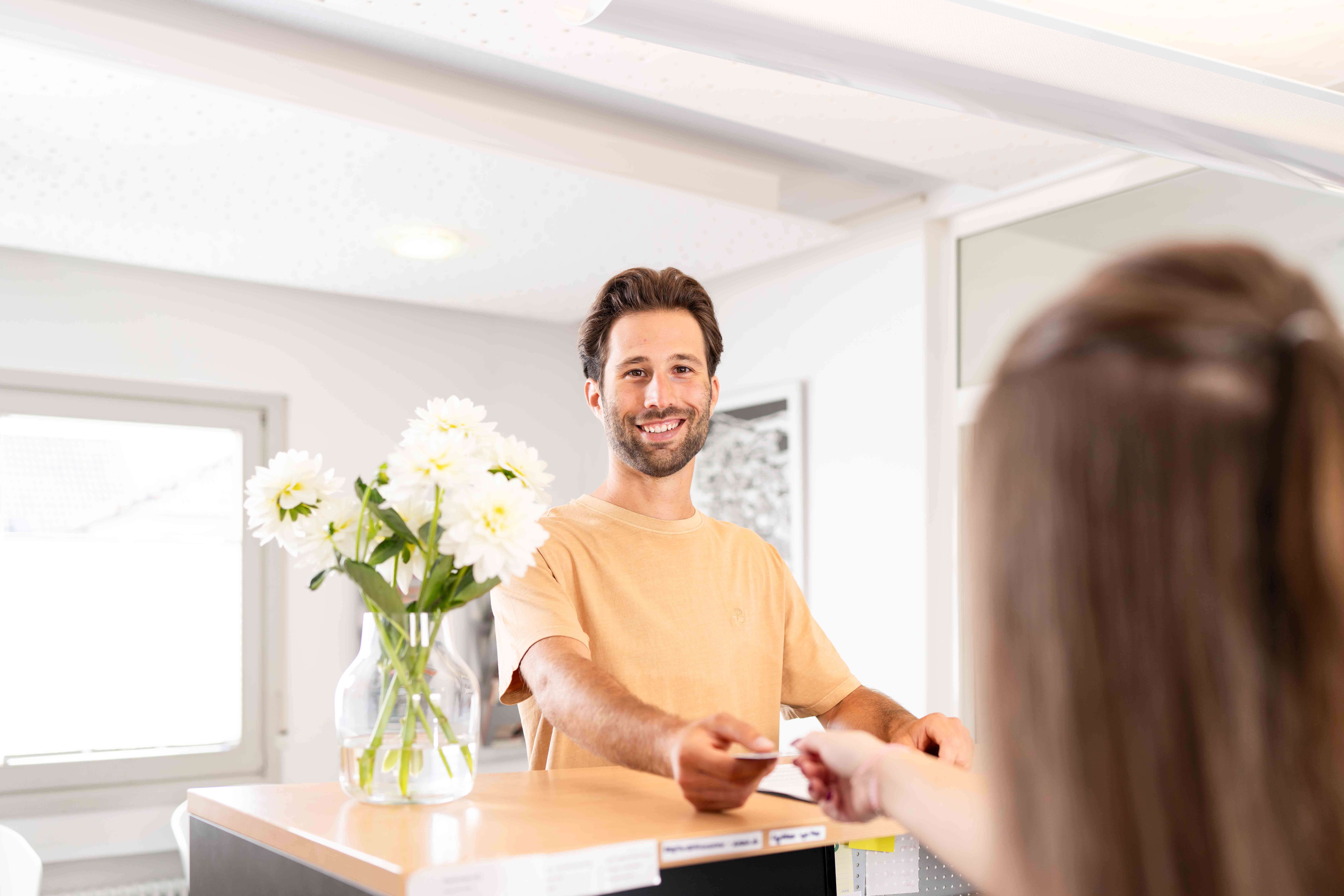 Smiling man handing a card to a woman at a reception desk with a vase of white flowers.