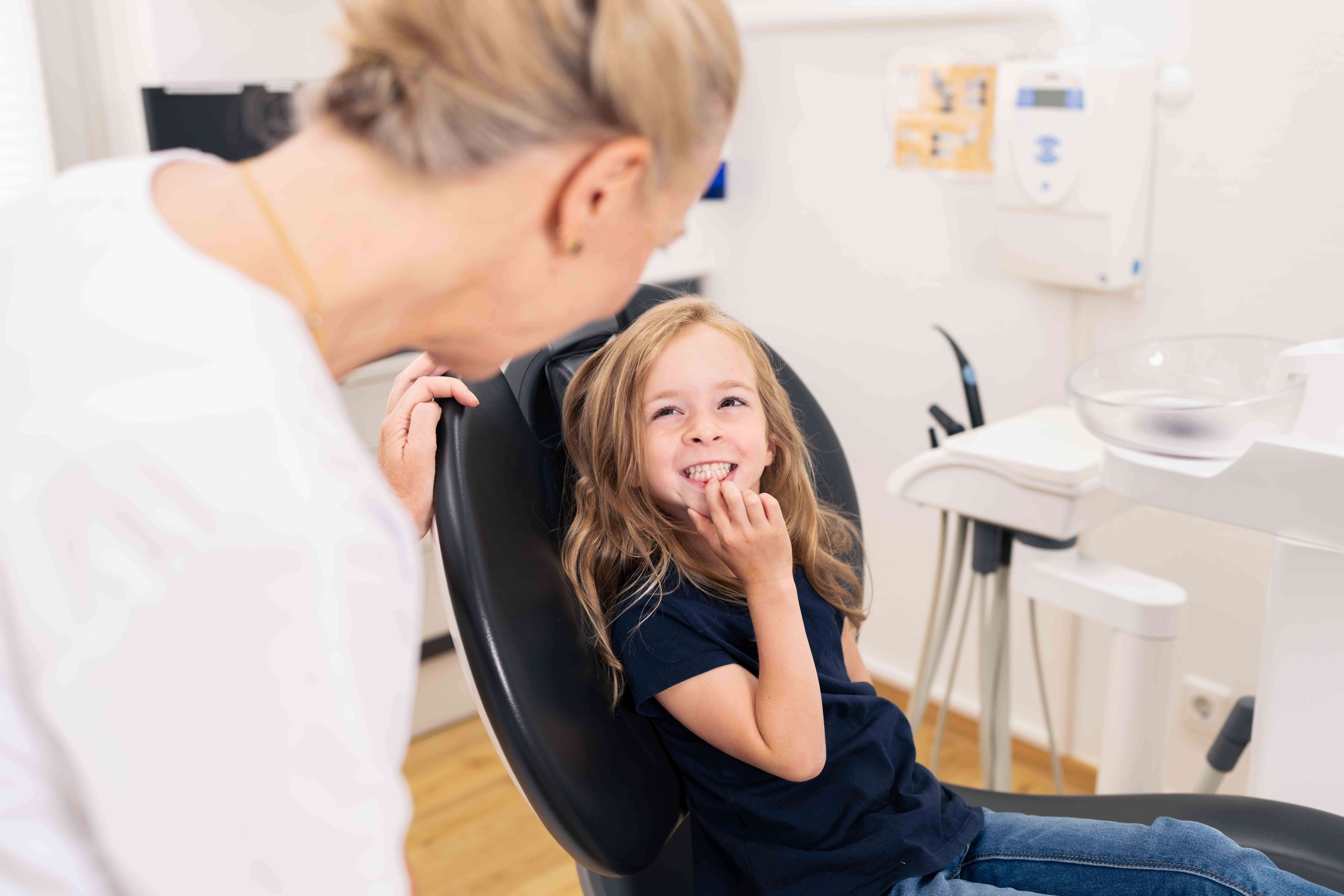 Young girl smiling and showing her teeth while sitting in a dental chair, with a dentist leaning towards her.
