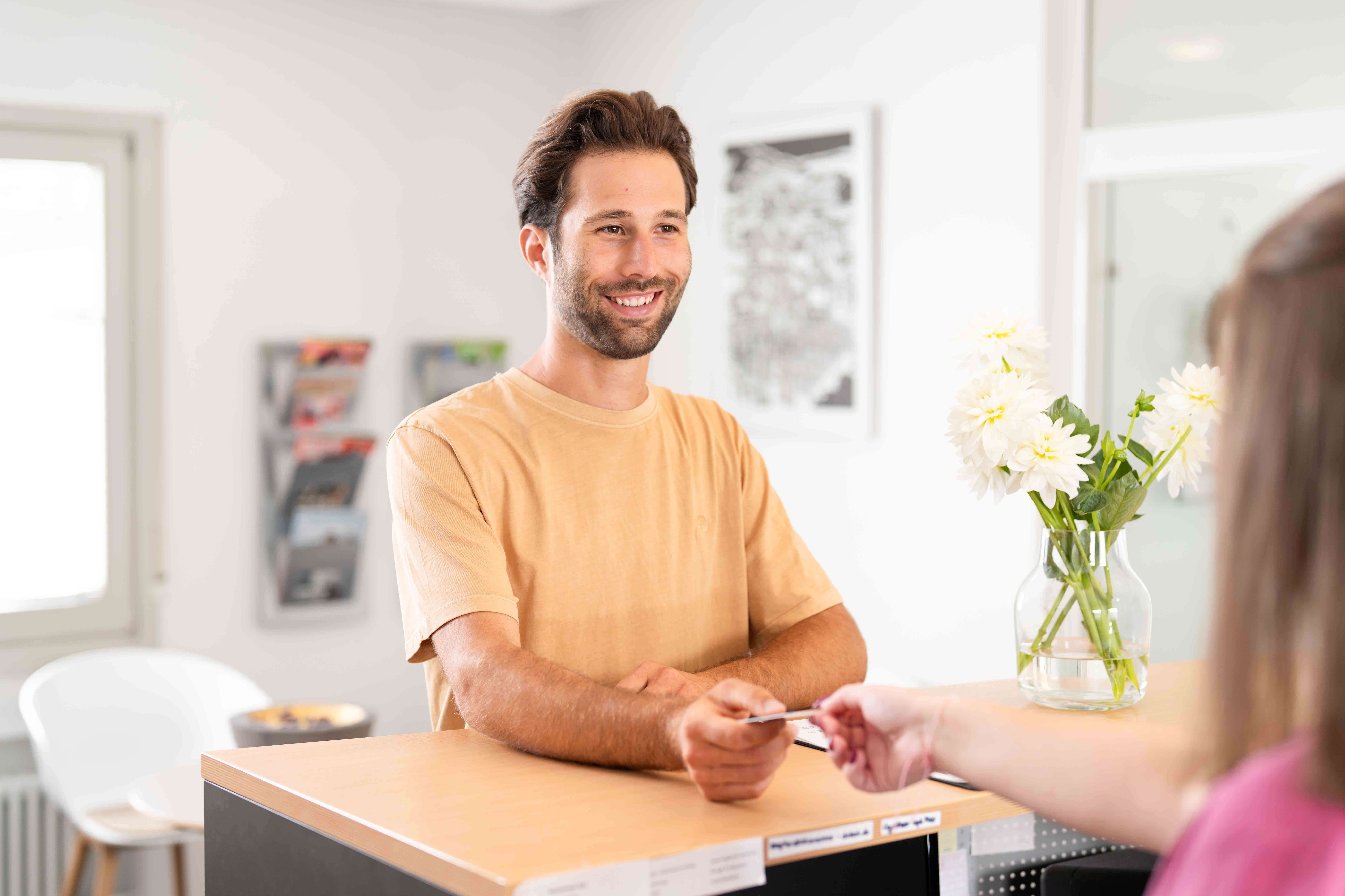 Smiling man in a beige t-shirt receiving a card from a person at a reception desk with white flowers in a vase.