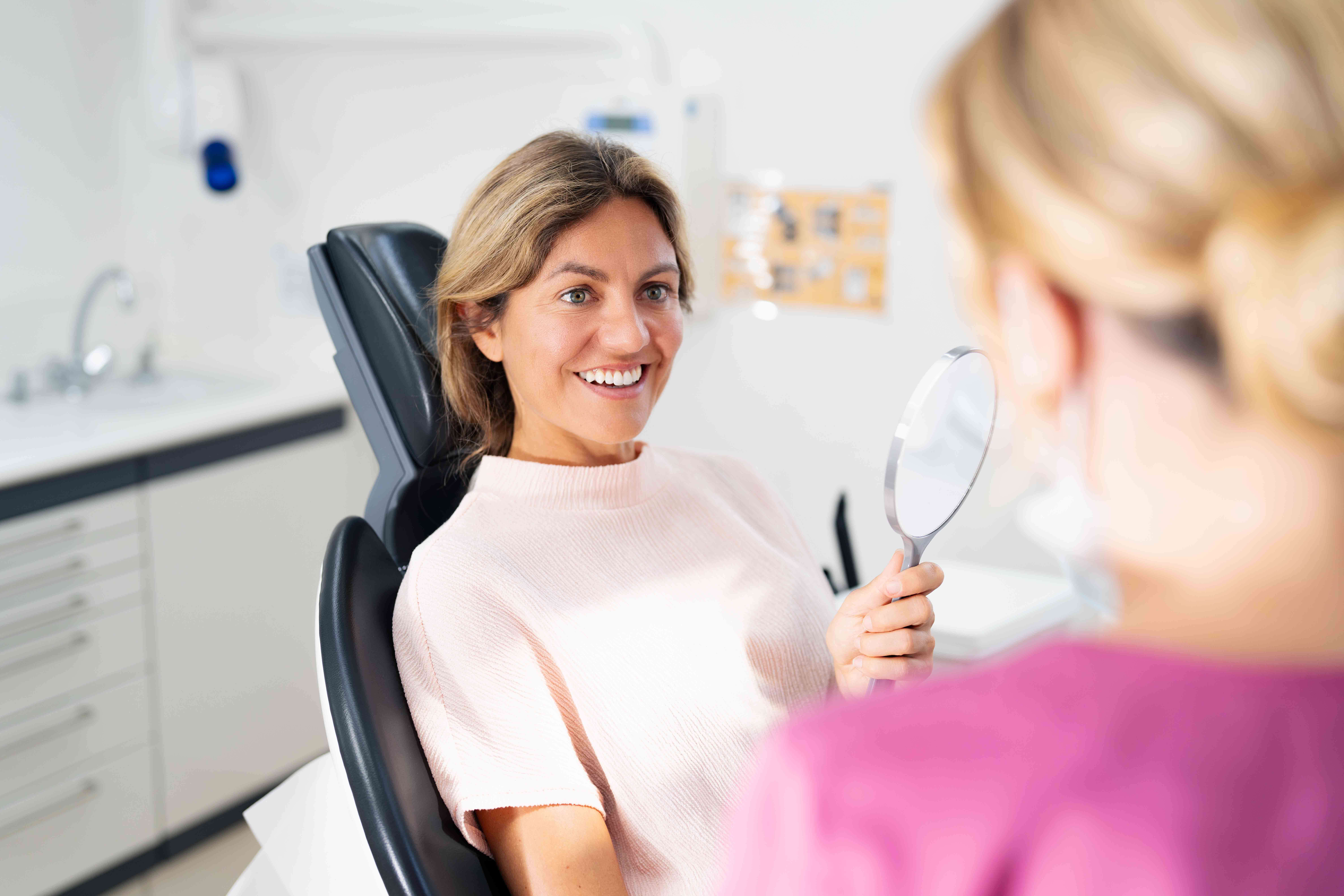 Smiling woman sitting in a dental chair holding a mirror, looking at a dental professional in a pink uniform.