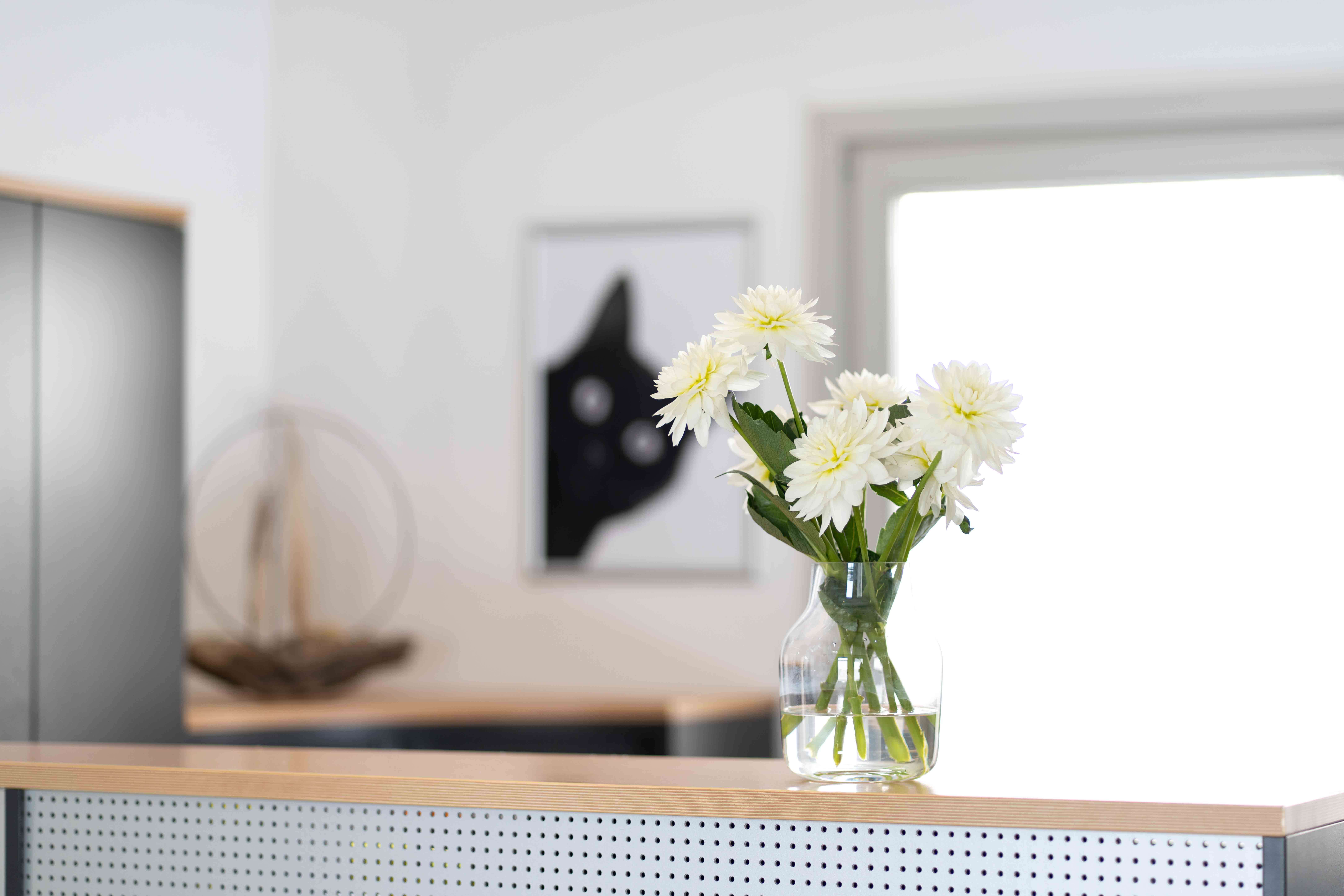 Clear glass vase with white flowers on a modern perforated desk with a blurred black cat artwork in the background.