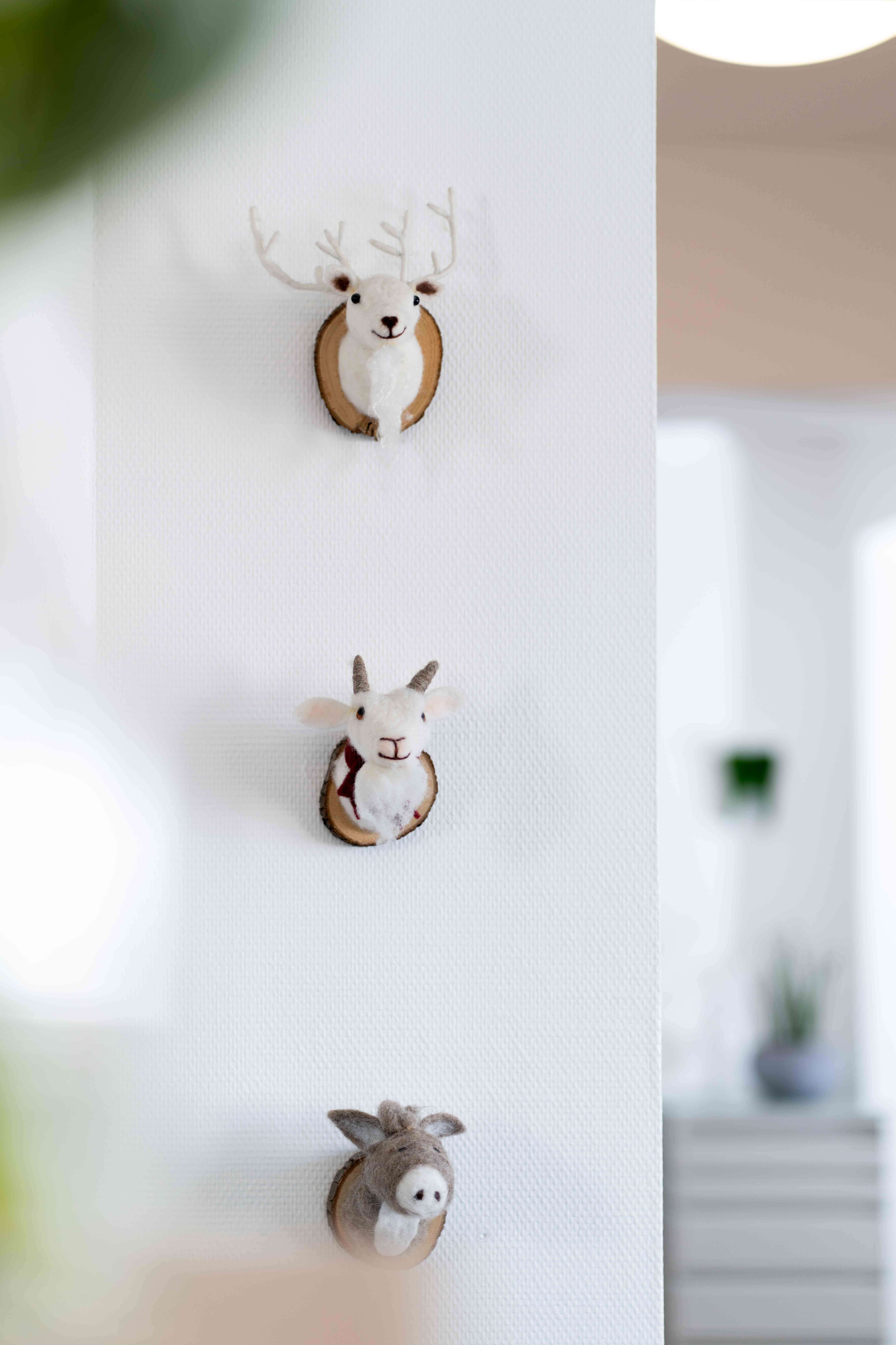 Three small felt animal head wall mounts—deer with antlers, goat, and donkey—on a white wall in a bright room.