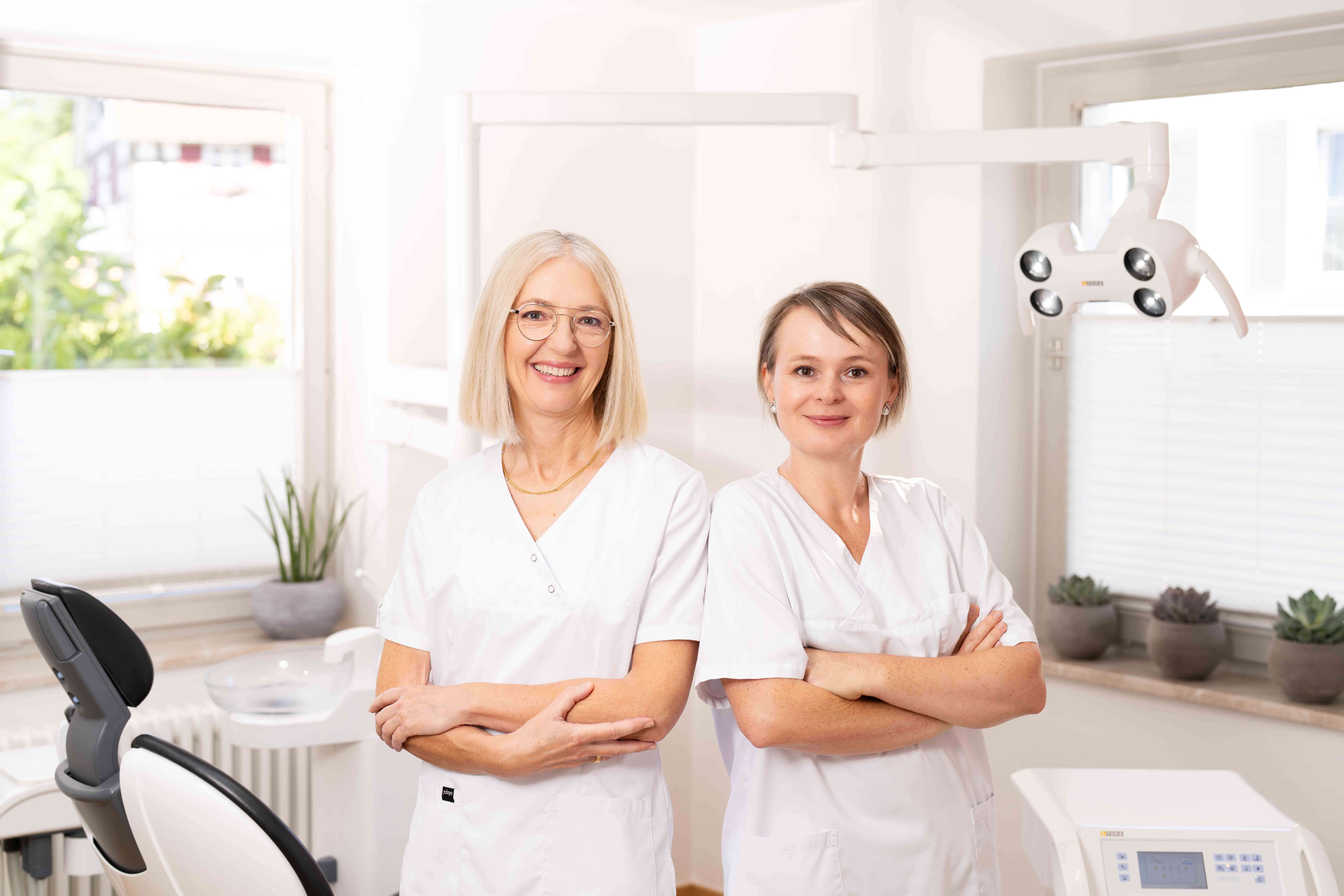 Two female dental professionals in white uniforms standing with arms crossed in a bright dental office.