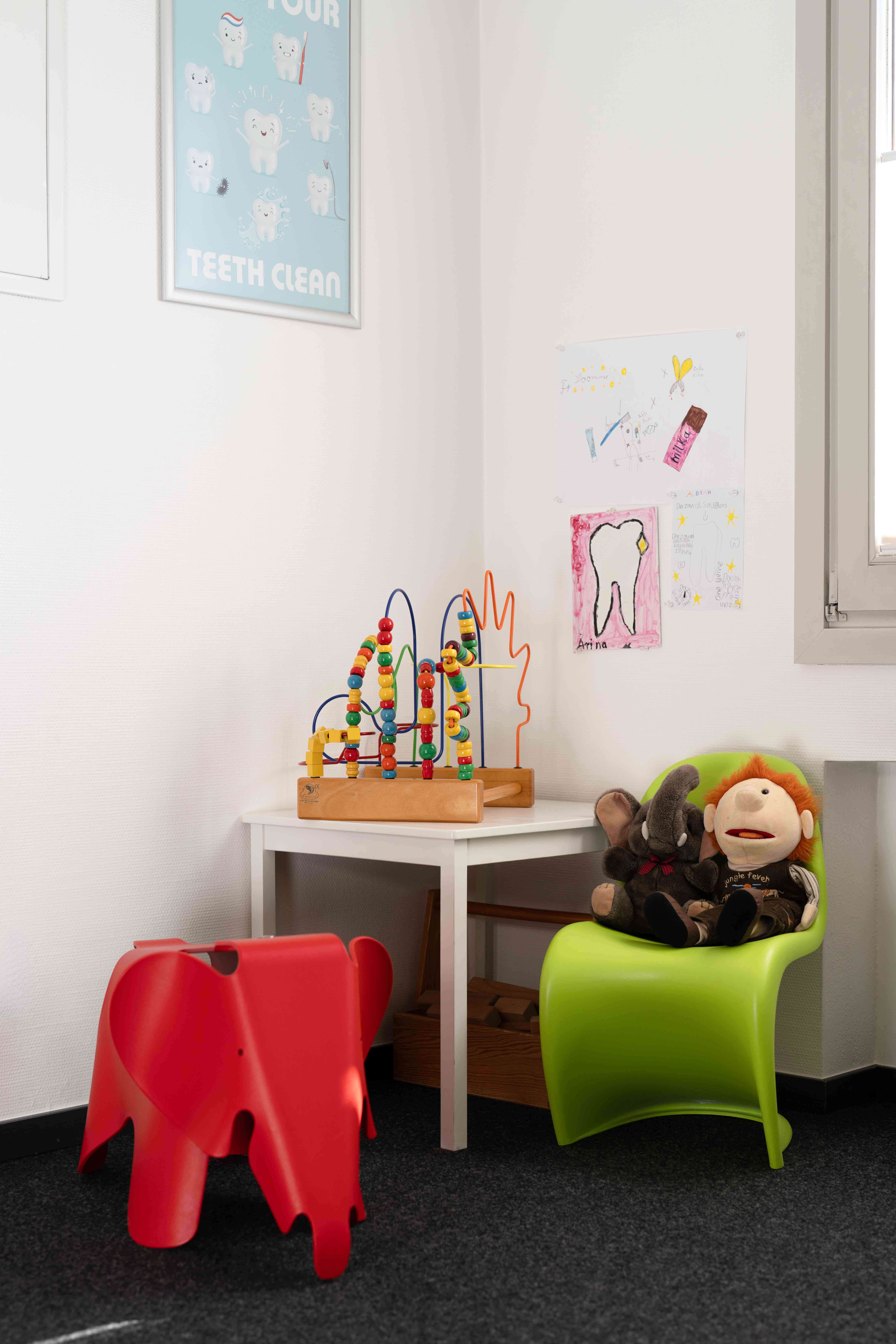 Children's play corner with a red elephant-shaped chair, a white table with a colorful bead maze toy, a green chair holding two stuffed animals, and children's drawings on the wall.