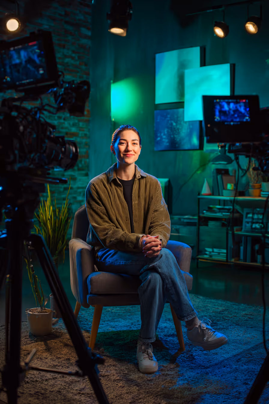 Smiling woman in casual clothes sitting on a chair in a colorful studio setup surrounded by cameras and lights.