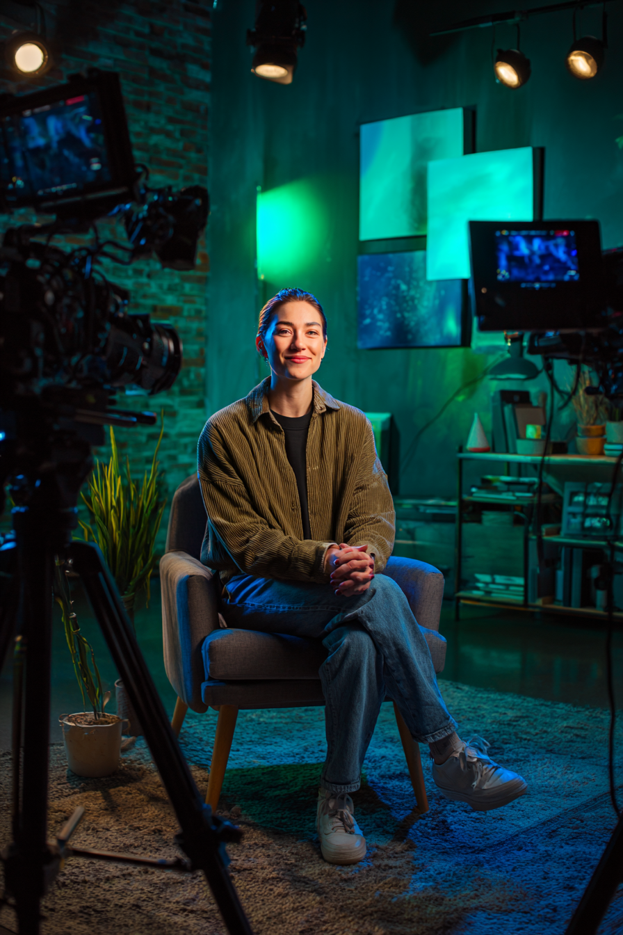 Smiling woman in casual clothes sitting on a chair in a colorful studio setup surrounded by cameras and lights.
