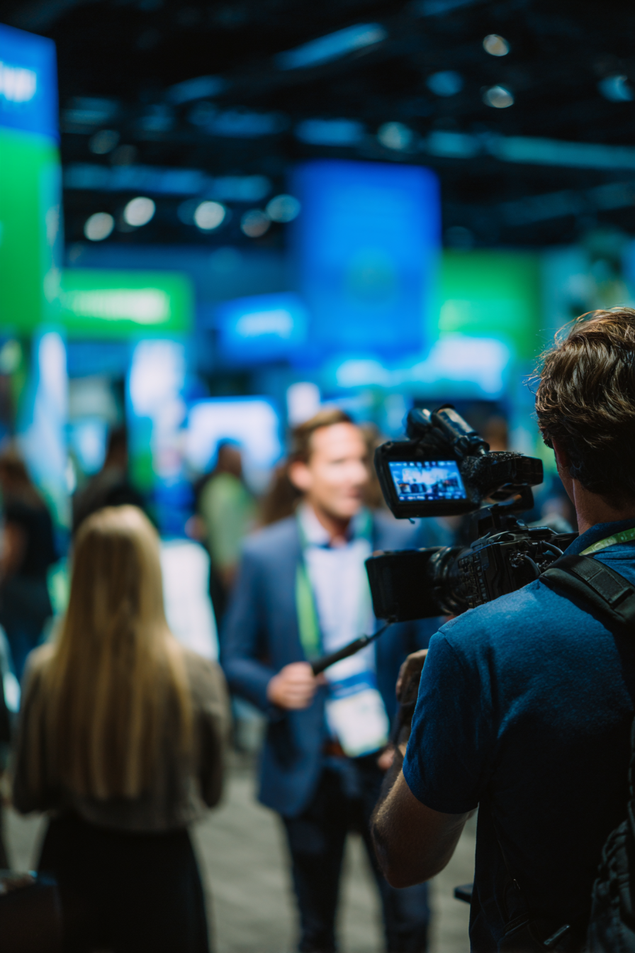 Camera operator filming a man in a suit at a busy indoor event with colorful blurred banners in the background.