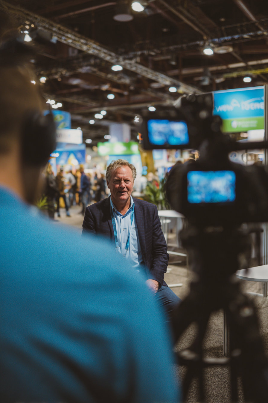 Man in a blue shirt and dark jacket sitting and talking in front of a video camera at an indoor event with crowd and booths in the background.