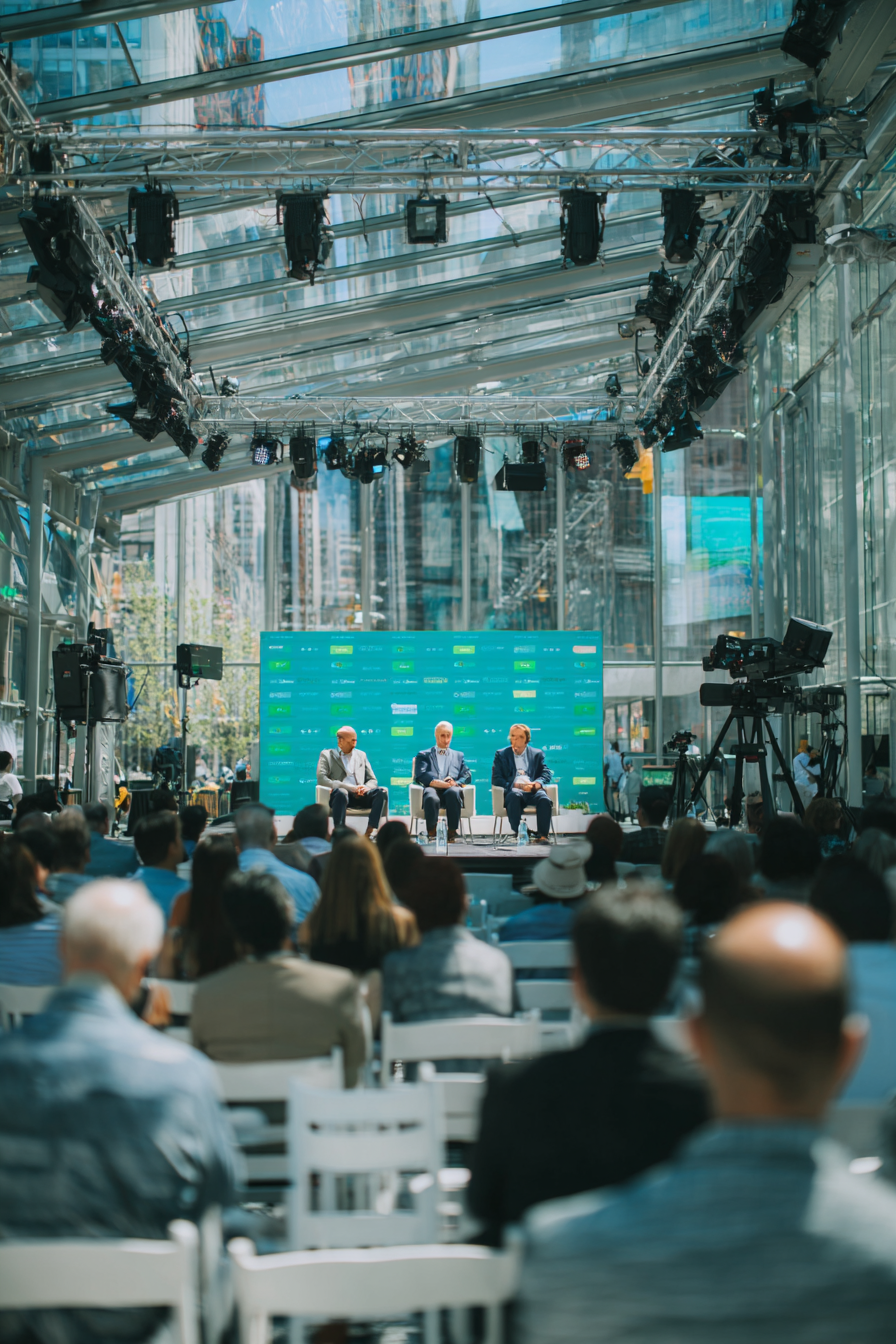 Three men seated on a stage with a turquoise backdrop, speaking to an audience in a glass-walled venue with professional lighting and cameras.