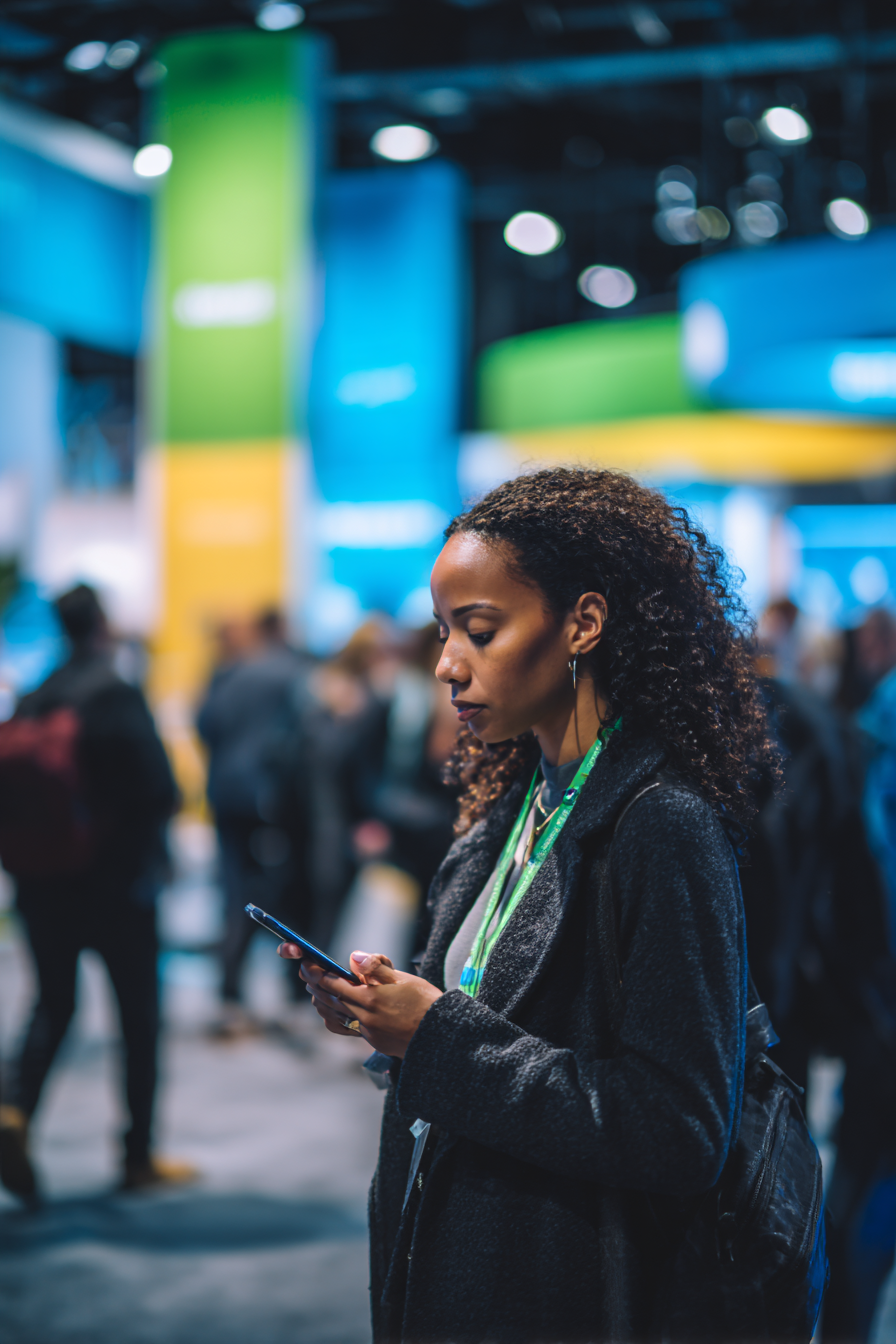 Woman with curly hair wearing a dark coat and green lanyard looking at her phone in a busy indoor event space.
